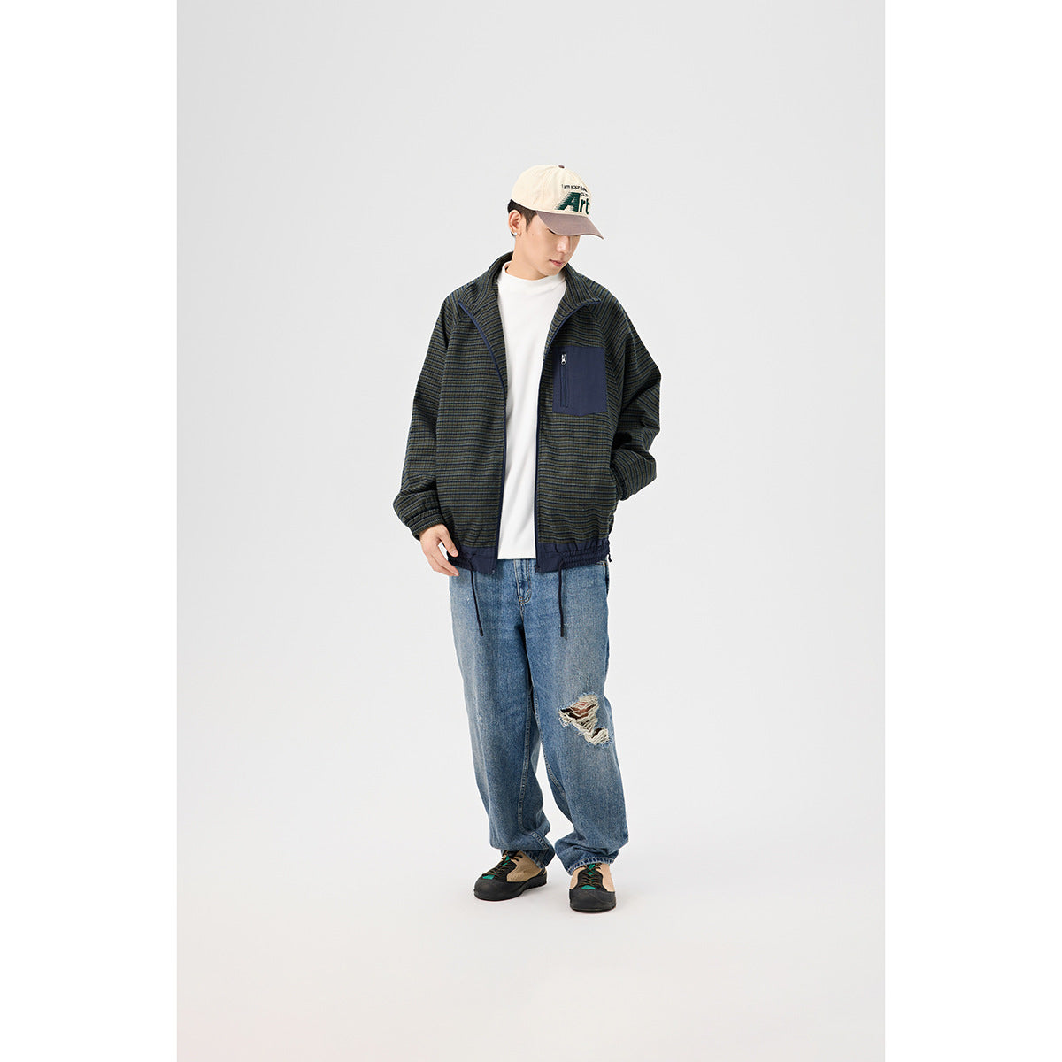 Full-length model wearing a dark striped zip jacket with a navy chest pocket, white shirt, loose jeans and cap.