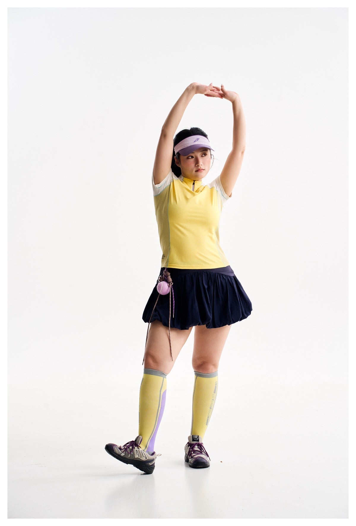 Model stretching in a yellow and ivory athletic top, navy bubble skirt, yellow socks and purple visor.