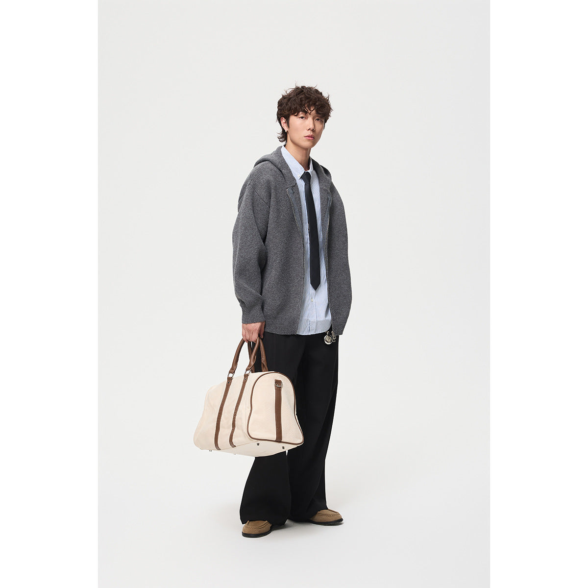 Model wearing a charcoal-gray hooded zip cardigan with a striped shirt and tie, holding a cream duffel bag.