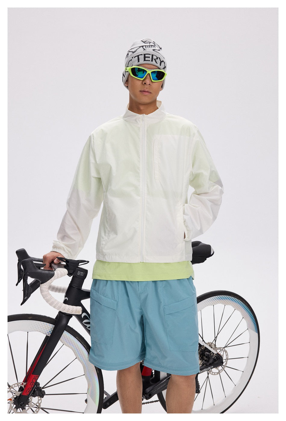 Front view of a man wearing a pale zip jacket over a lime shirt and blue shorts, with a road bike behind him.