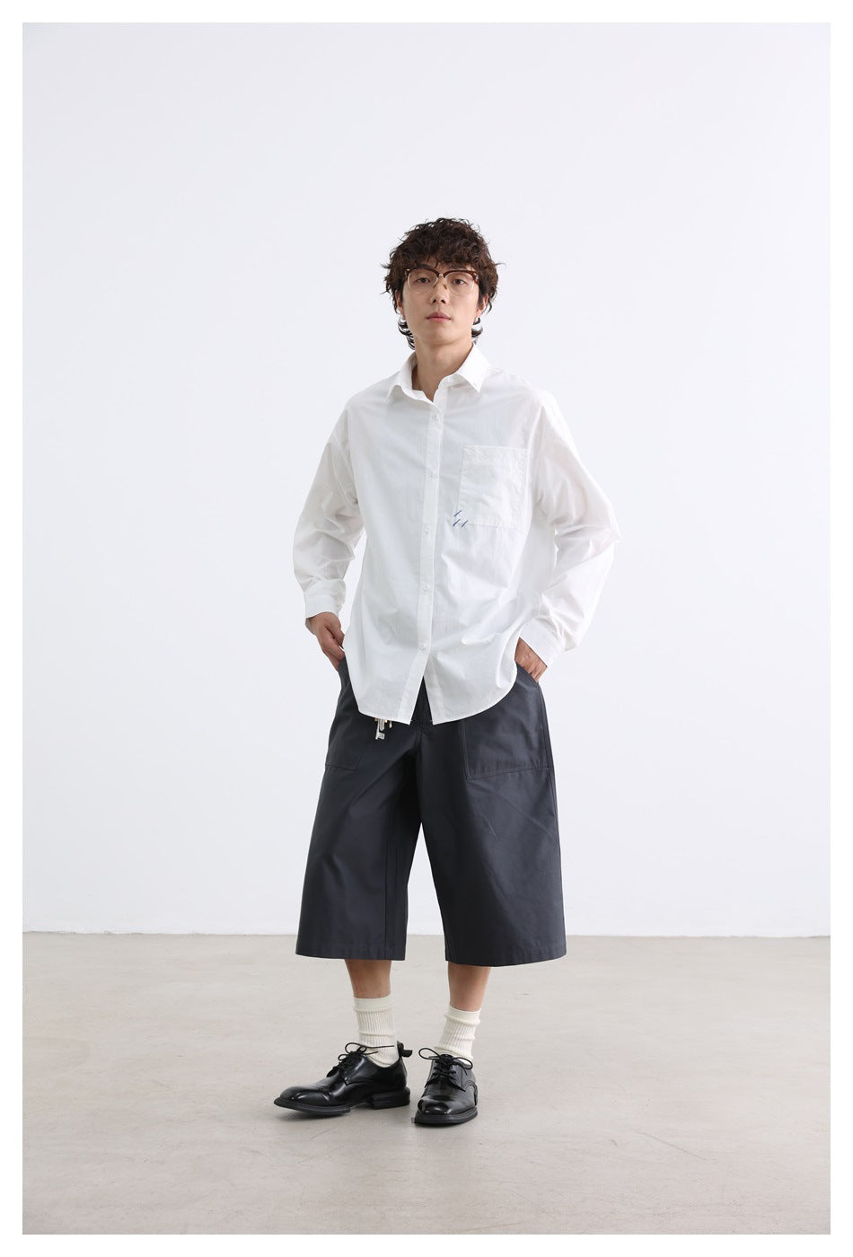 Full-length front view of a model wearing a white button-down shirt, charcoal knee-length shorts, and black shoes.