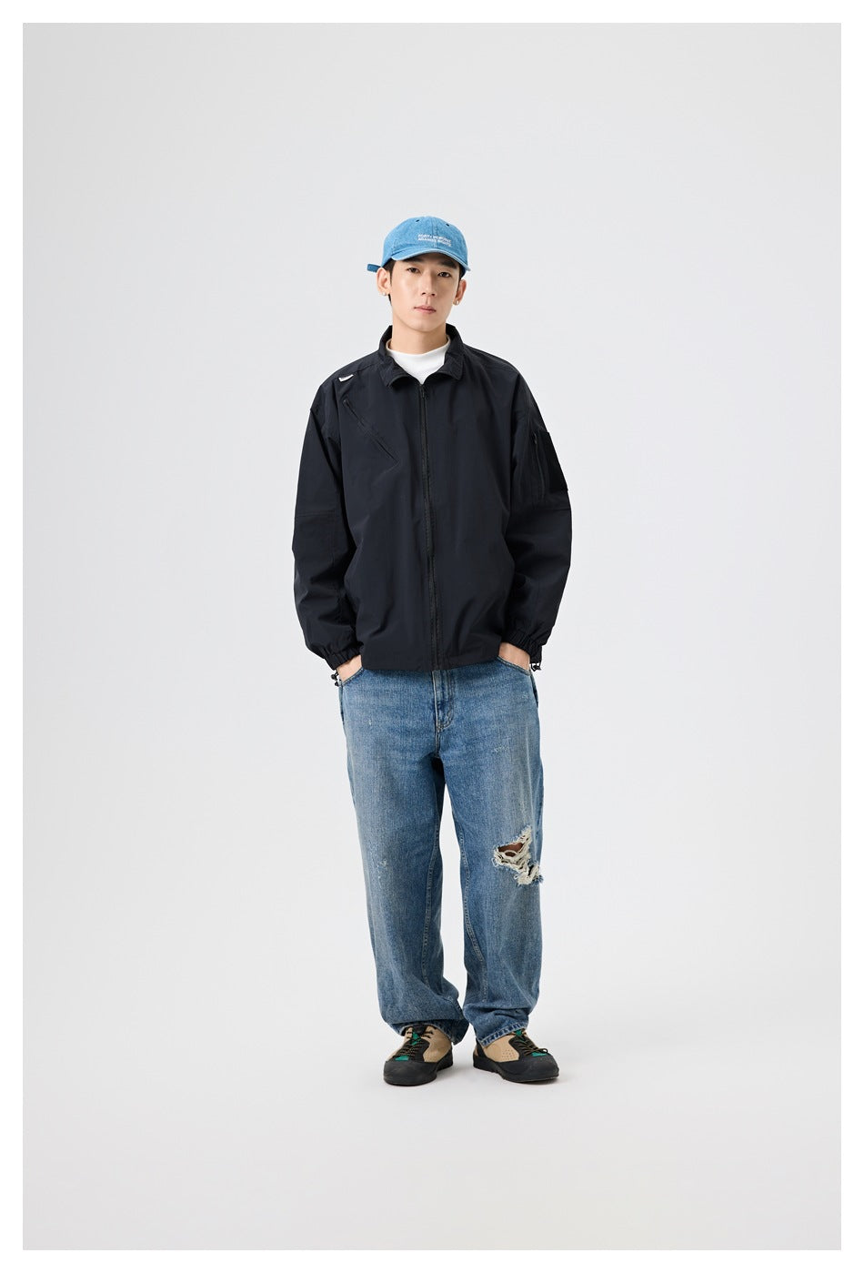 Full-length model view of a black zip-front jacket with elastic cuffs, styled with jeans and a blue cap.