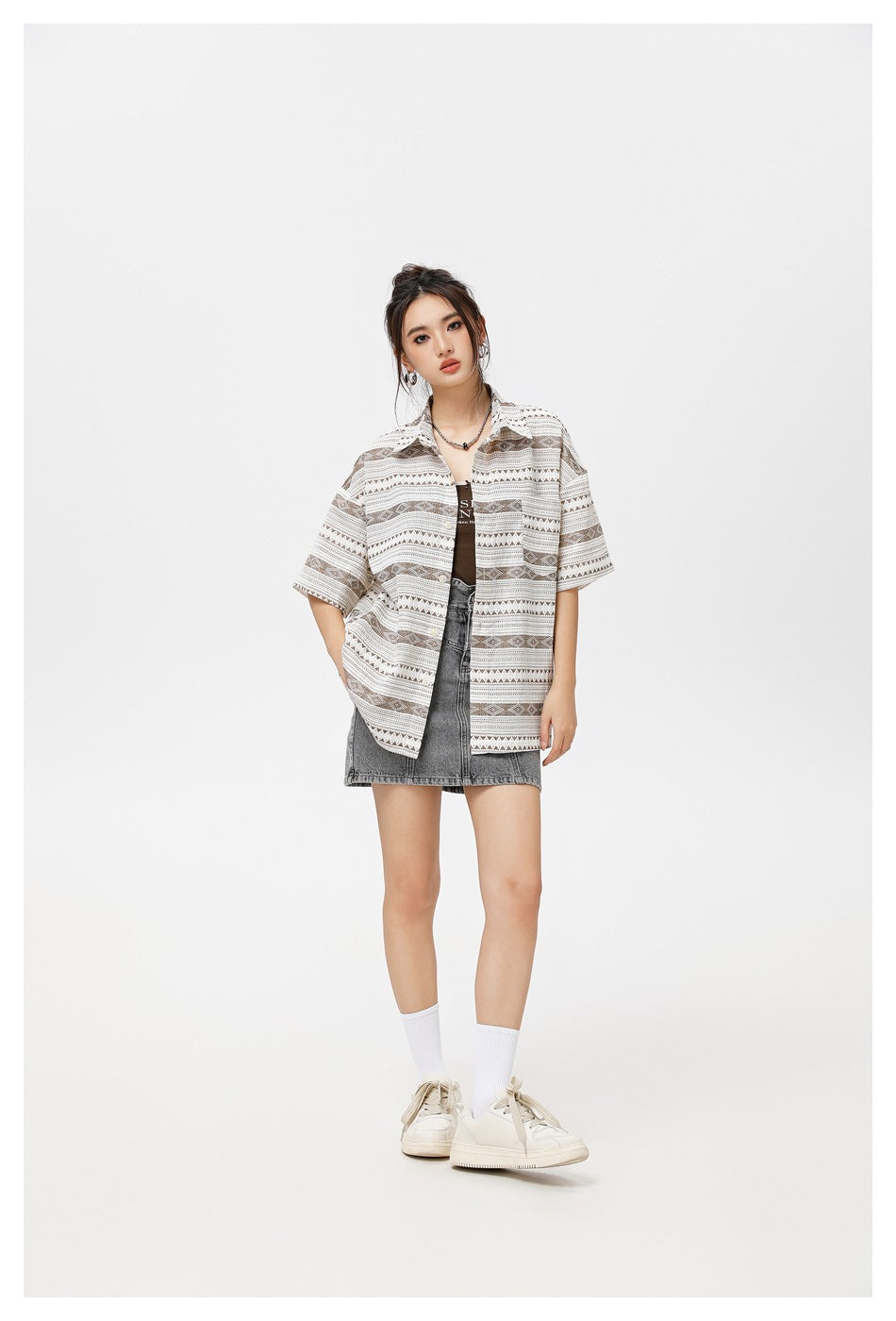 Model in the brown geometric striped short-sleeve shirt worn open with a denim skirt and white sneakers