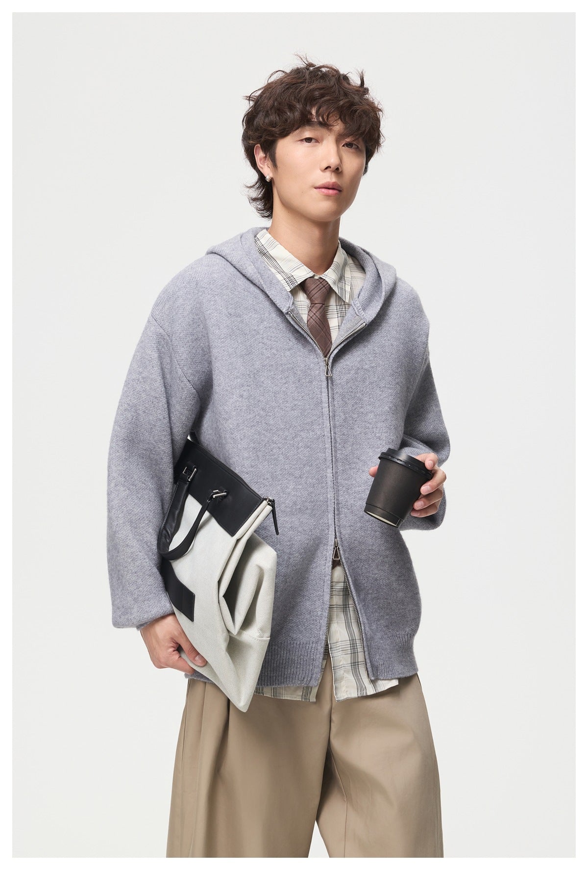 Model wearing a light gray zip-up hooded knit jacket, holding a black-and-white bag and a cup.