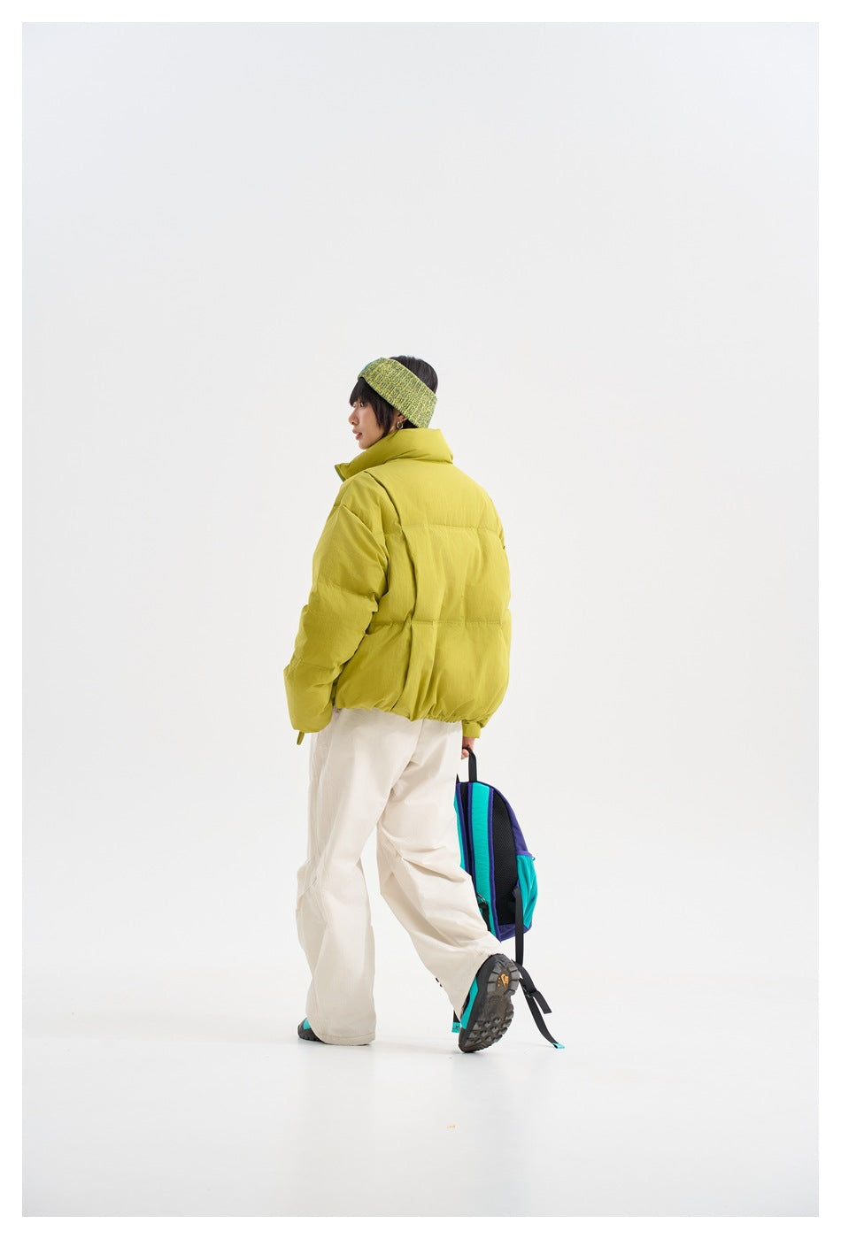 Rear three-quarter view of a model wearing a lime-yellow puffer jacket and white trousers.