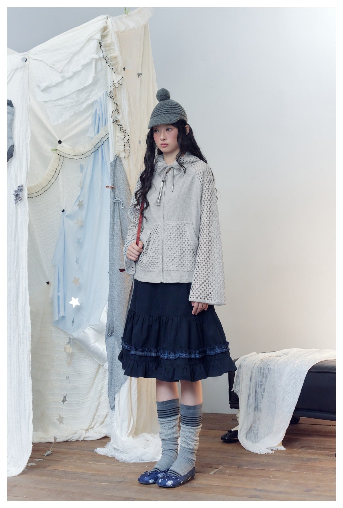 Model in gray mesh-sleeve zip hooded jacket with navy ruffled skirt, three-quarter view showing hood drawstring and pocket