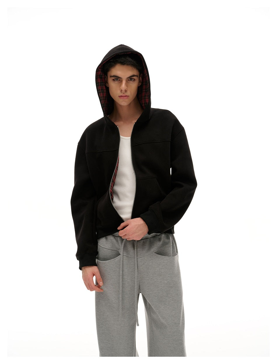 Model wearing a black zip-up hoodie with the hood up and red plaid lining, paired with loose gray pants.