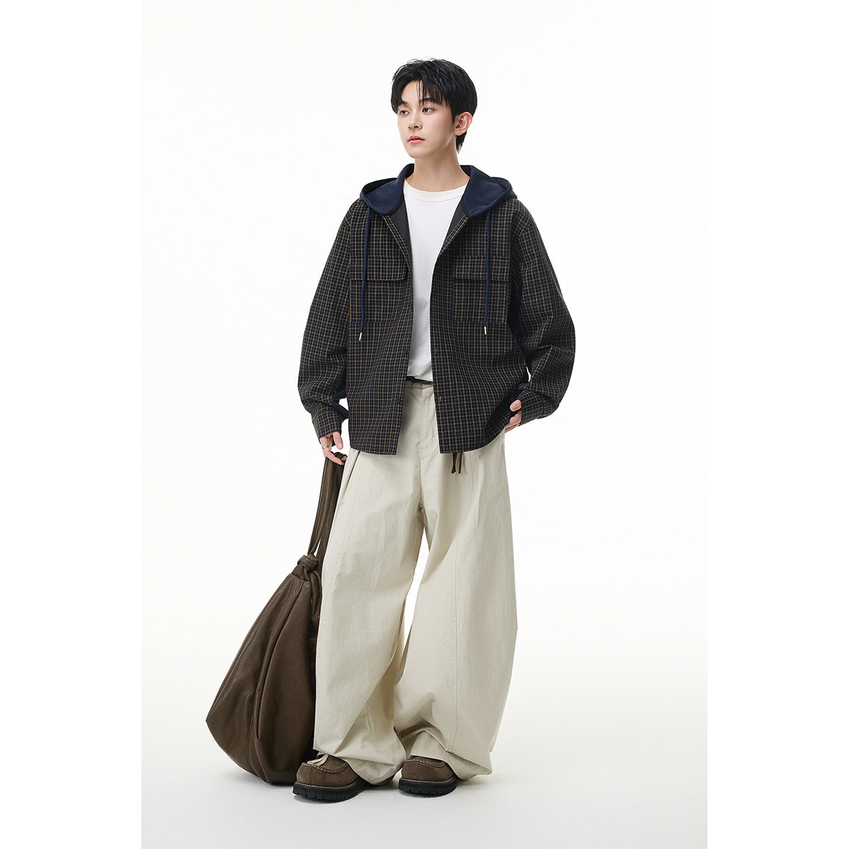 Full-length model wearing a dark plaid hooded shirt jacket with navy hood and details, white tee, and loose cream trousers.