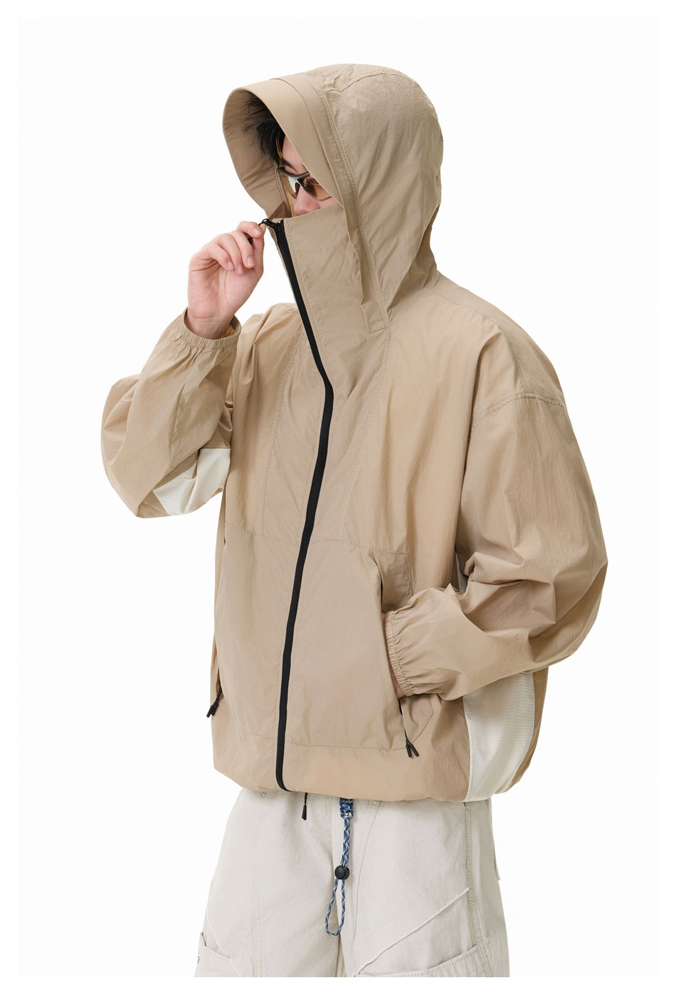 Side view of a model wearing a beige hooded jacket zipped over the face, with sunglasses and hands at the zipper.