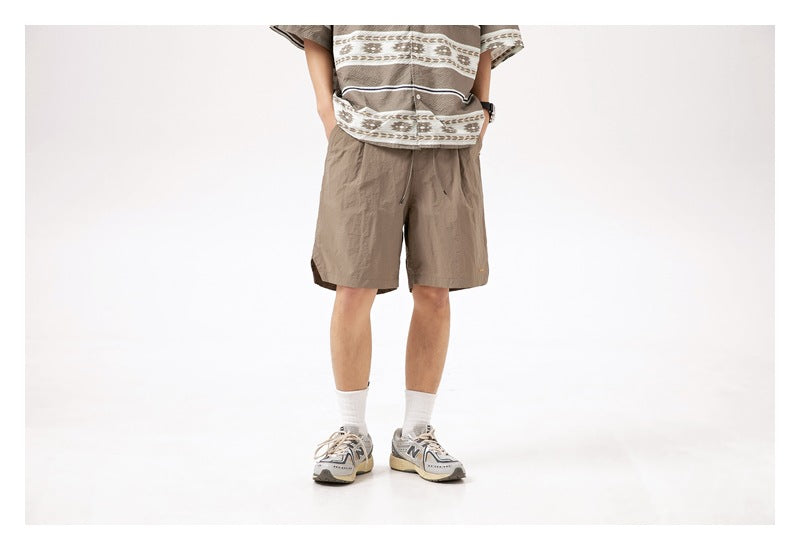 Cropped view of a model wearing taupe drawstring shorts beneath a patterned short-sleeve shirt.