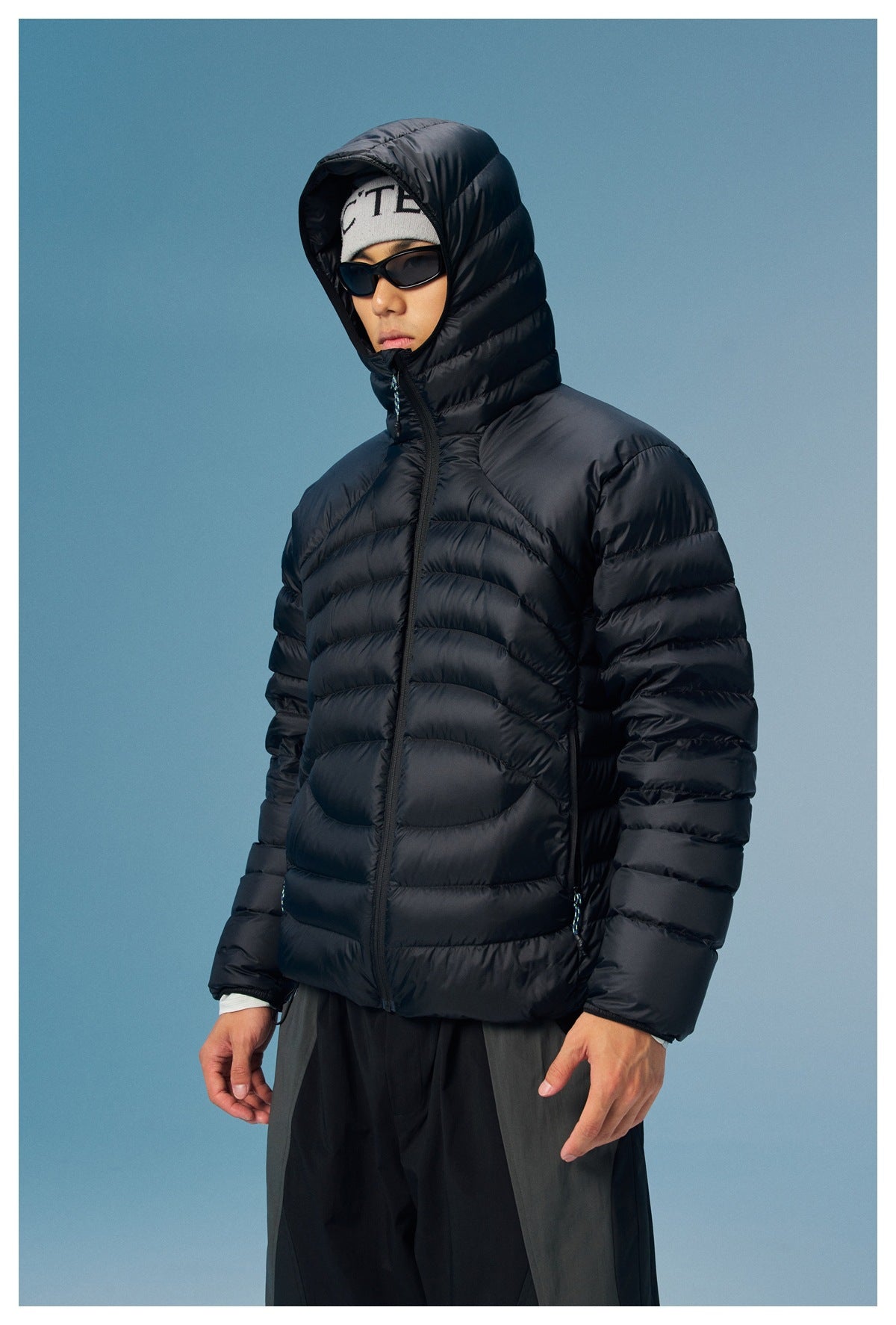 Model wearing a black quilted hooded zip jacket with horizontal baffles, shown from the front against a blue background.