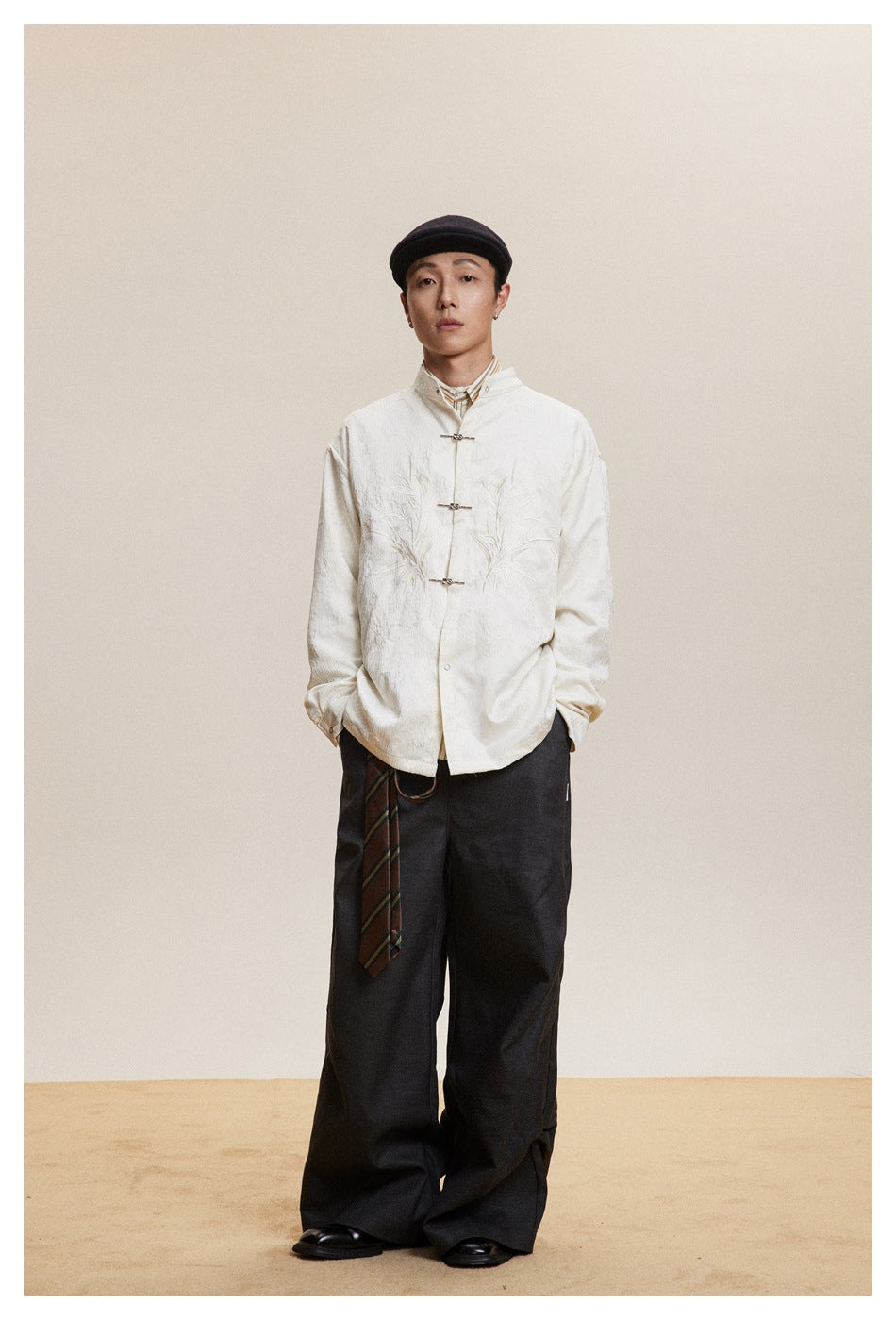 Model wearing the off-white embroidered stand-collar shirt with metal closures, dark wide trousers and a flat cap, front view