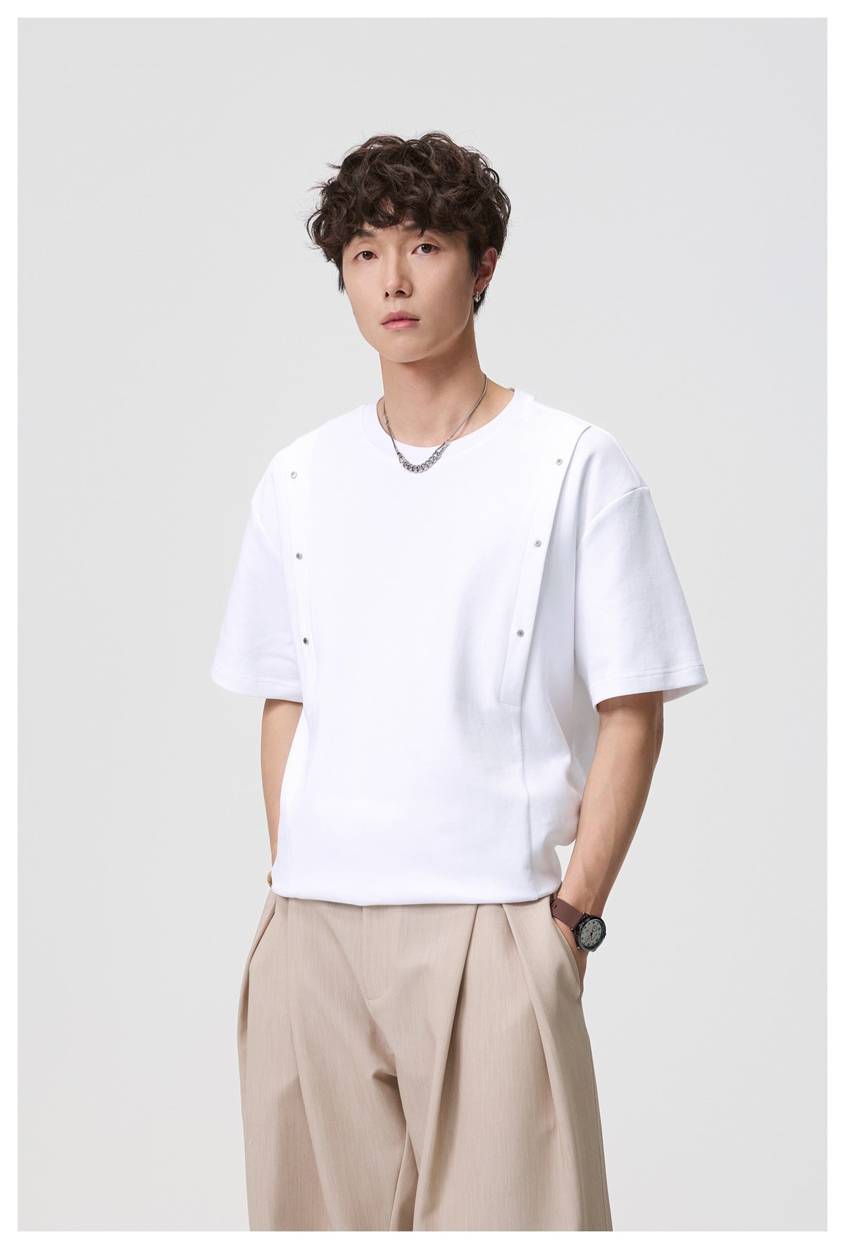 Model wearing an oversized white crew-neck tee with short sleeves and snap-fastened vertical front straps, paired with beige