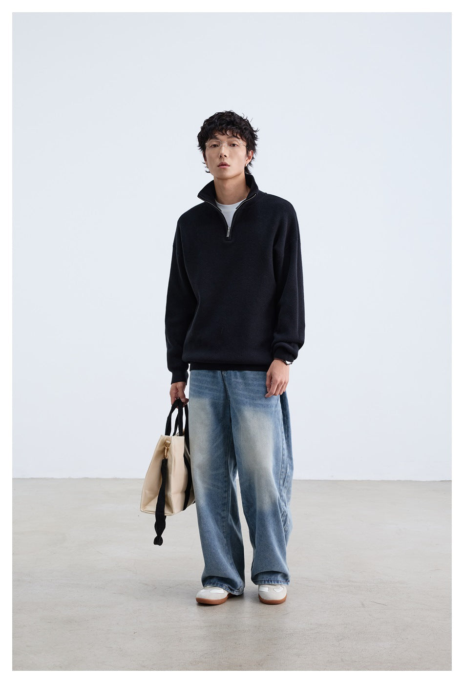 Full-length model wearing a black quarter-zip sweater, wide-leg blue jeans, and carrying a beige tote.
