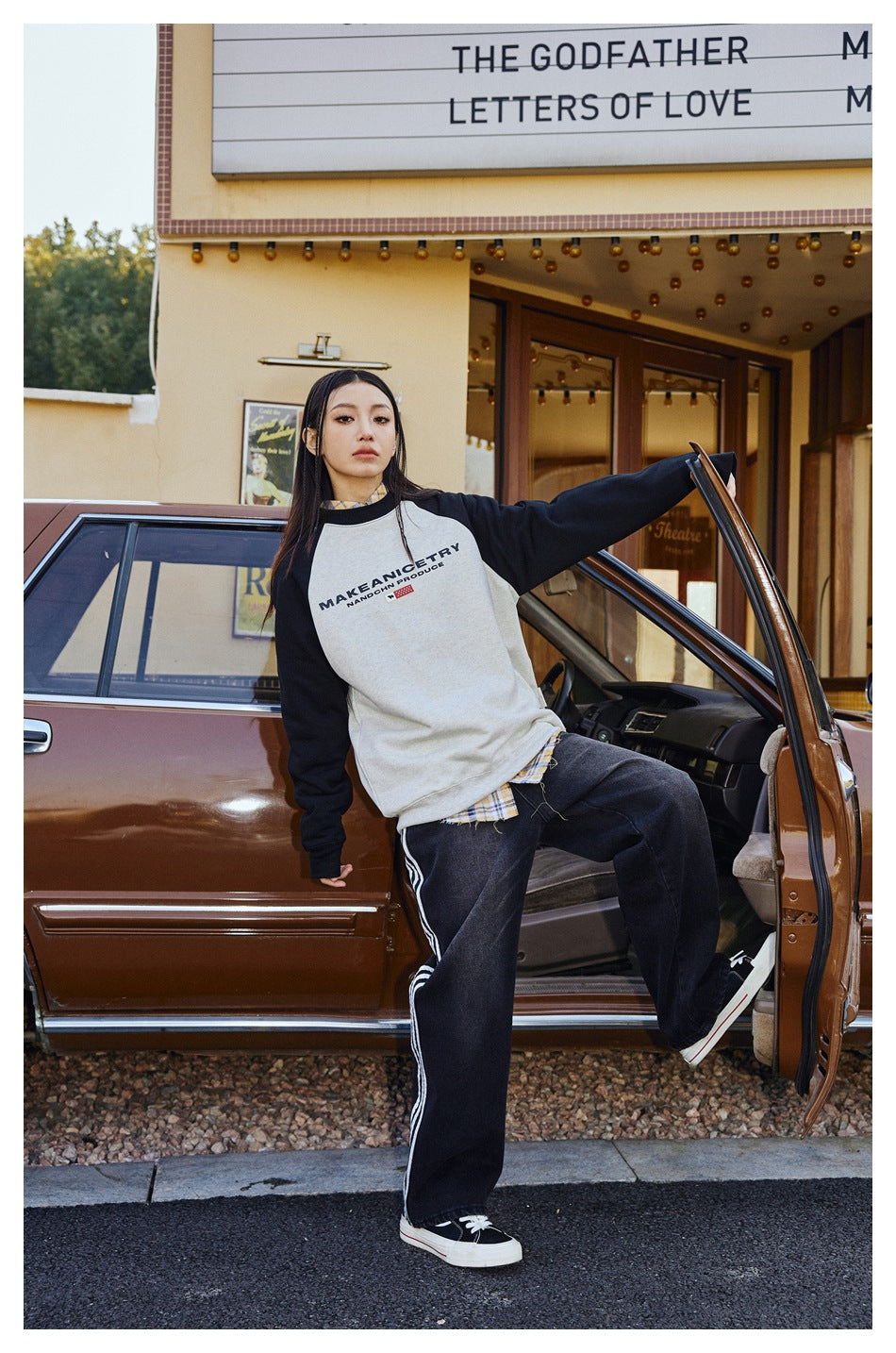 A model wears a cream raglan sweatshirt with black sleeves and dark striped pants beside a car.