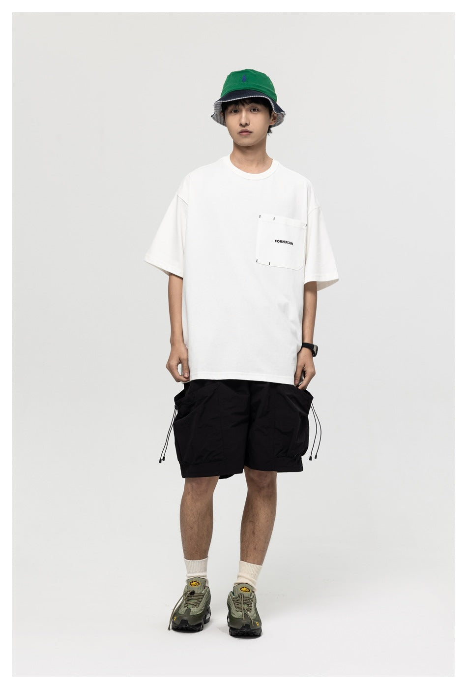 Model wearing a white relaxed pocket T-shirt with black shorts, bucket hat and green sneakers, standing front view
