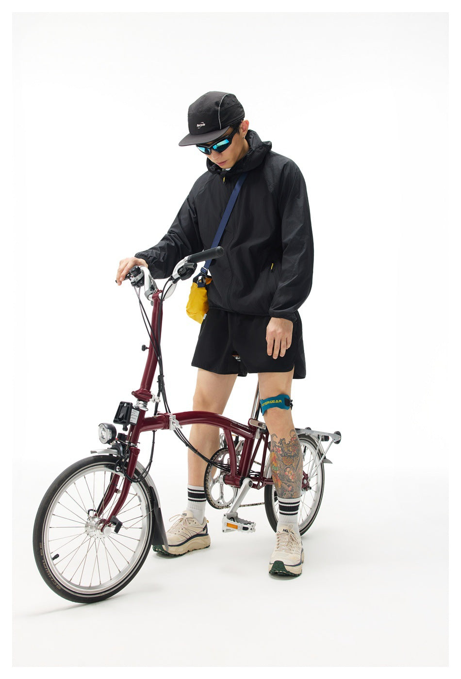 Person wearing a black hooded zip jacket with black shorts and a cap while standing beside a folding bicycle