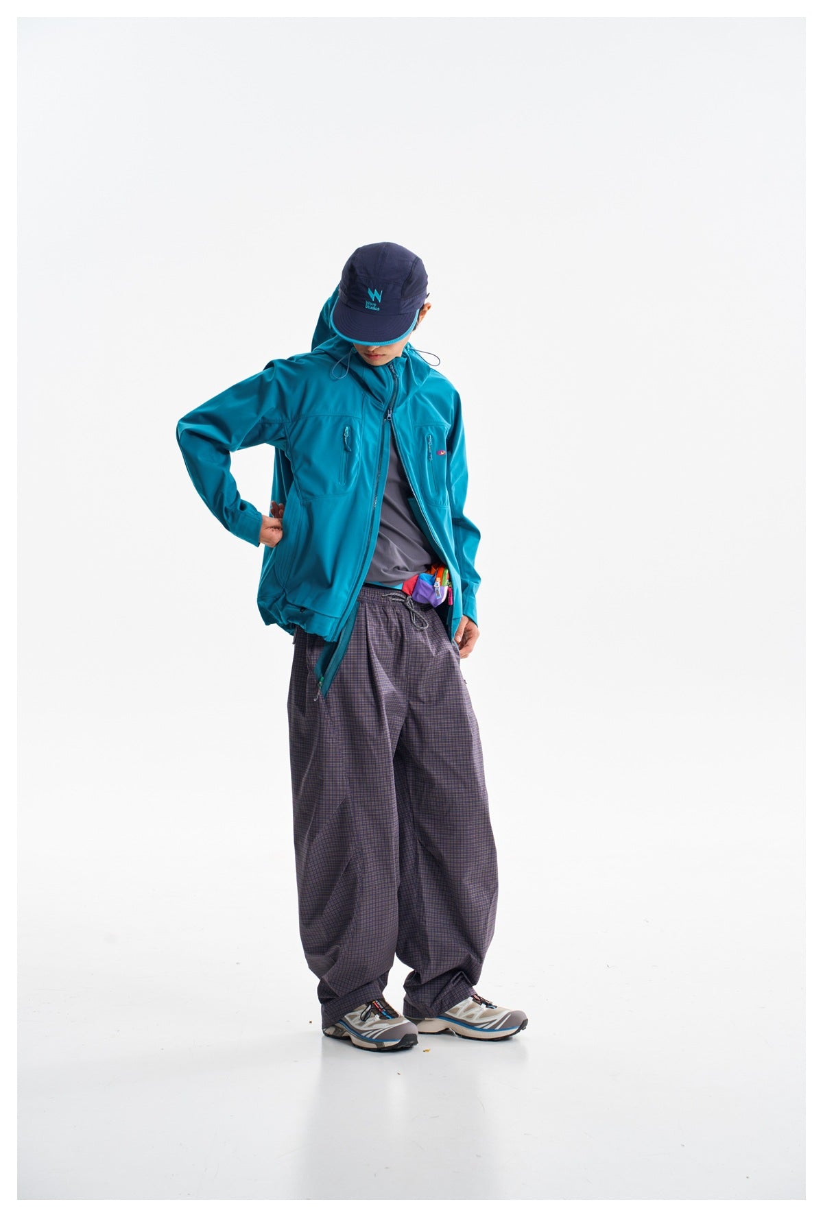 Model wearing a bright teal hooded jacket with zip pockets and loose dark plaid pants.