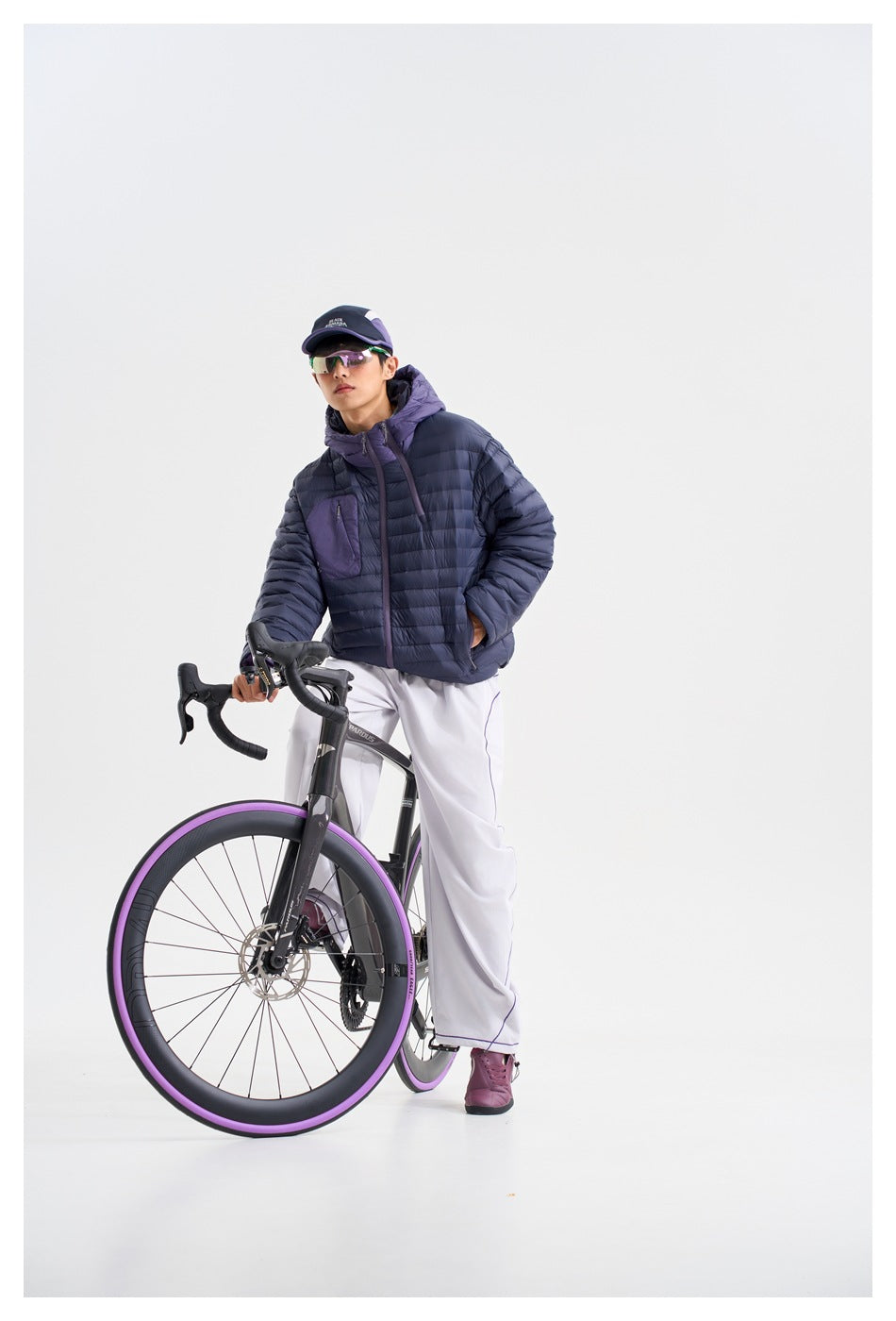 A person in a navy and purple hooded quilted jacket stands beside a road bicycle with purple wheel rims.