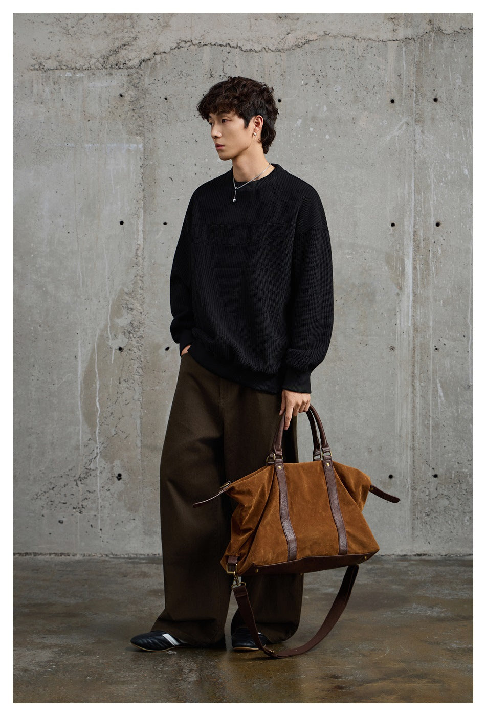 Model wearing an oversized black textured crewneck sweatshirt with subtle chest lettering and dark wide-leg trousers.