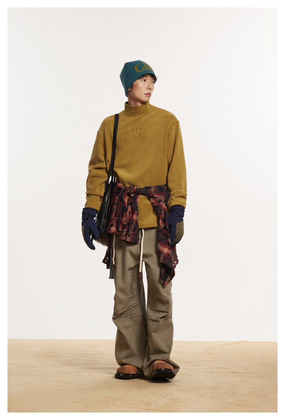 Full-length model wearing a mustard high-neck pullover, teal beanie, olive cargo pants, gloves, and a plaid shirt tied at the