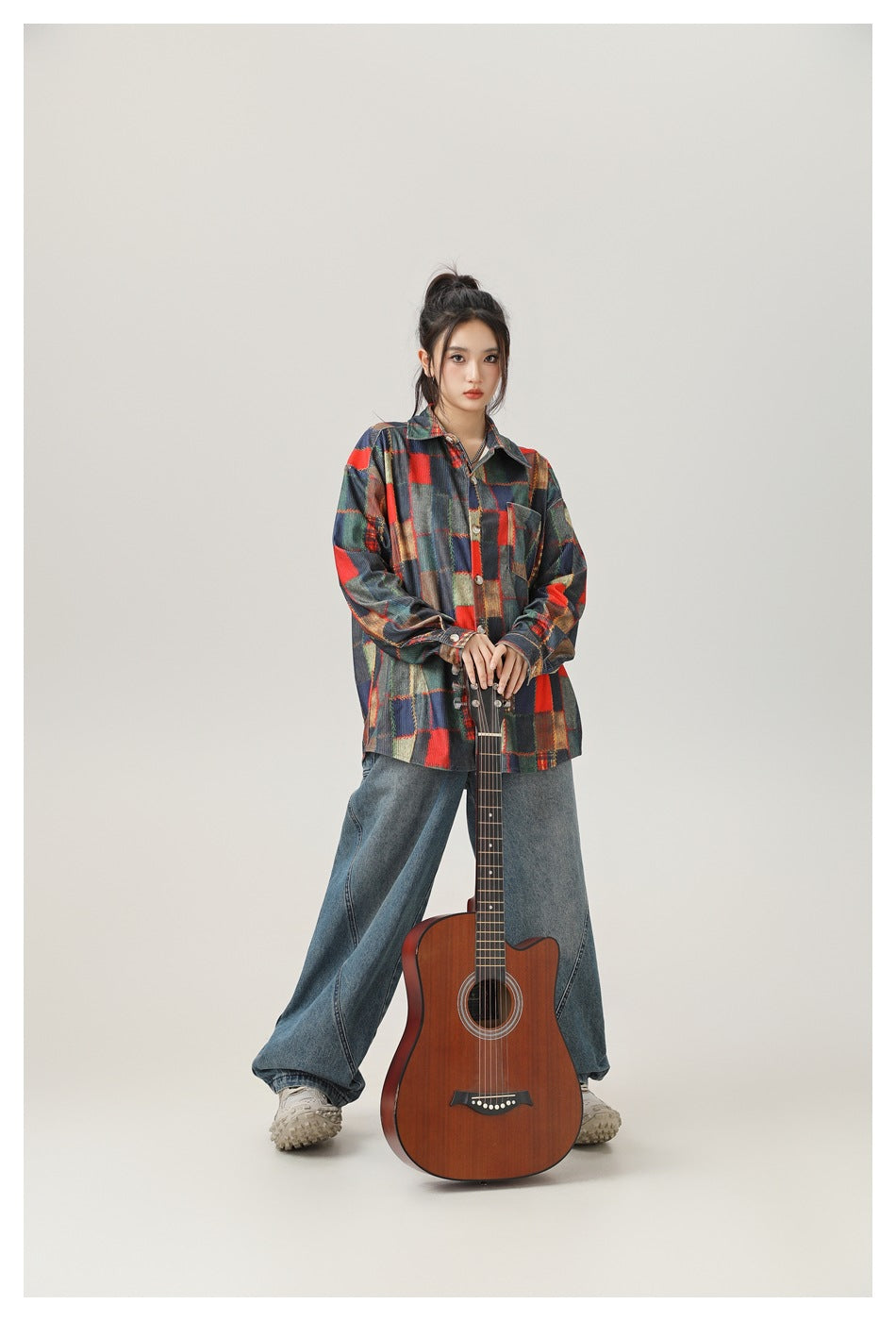 Model wearing a multicolor patchwork plaid button-up shirt and wide-leg jeans, standing behind a brown acoustic guitar.