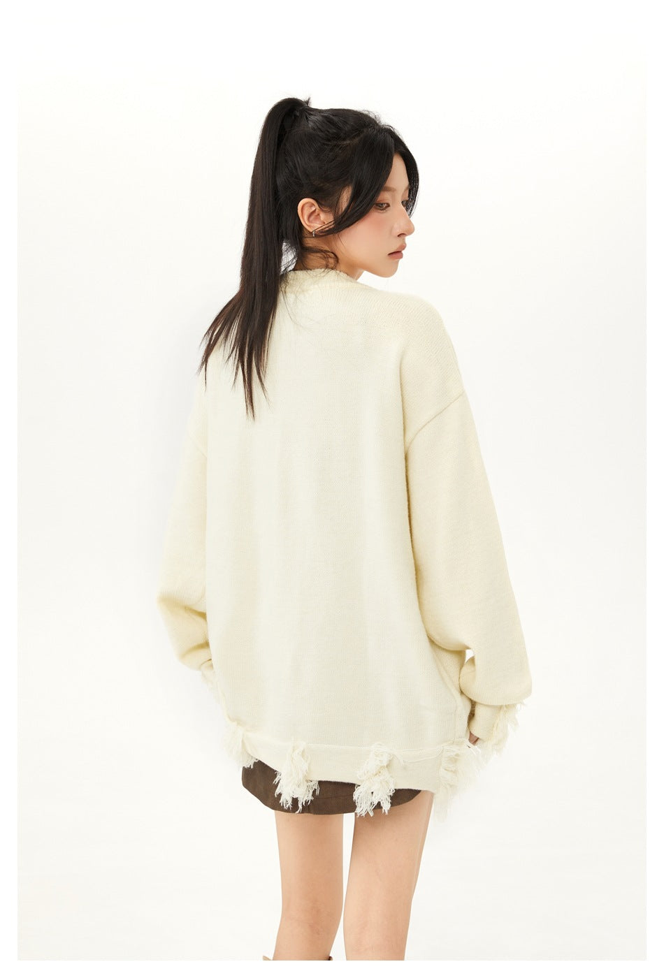 Model wearing an oversized cream sweater with fringe trim, shown from behind with a brown skirt.