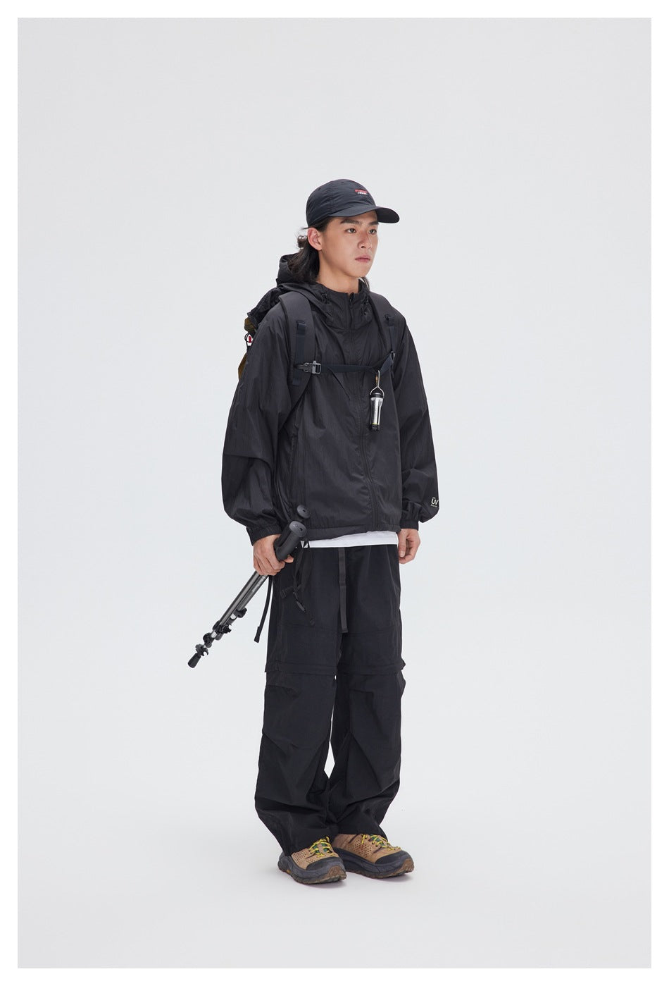Full-length model wearing a black hooded jacket and loose black trousers with a cap and hiking gear.