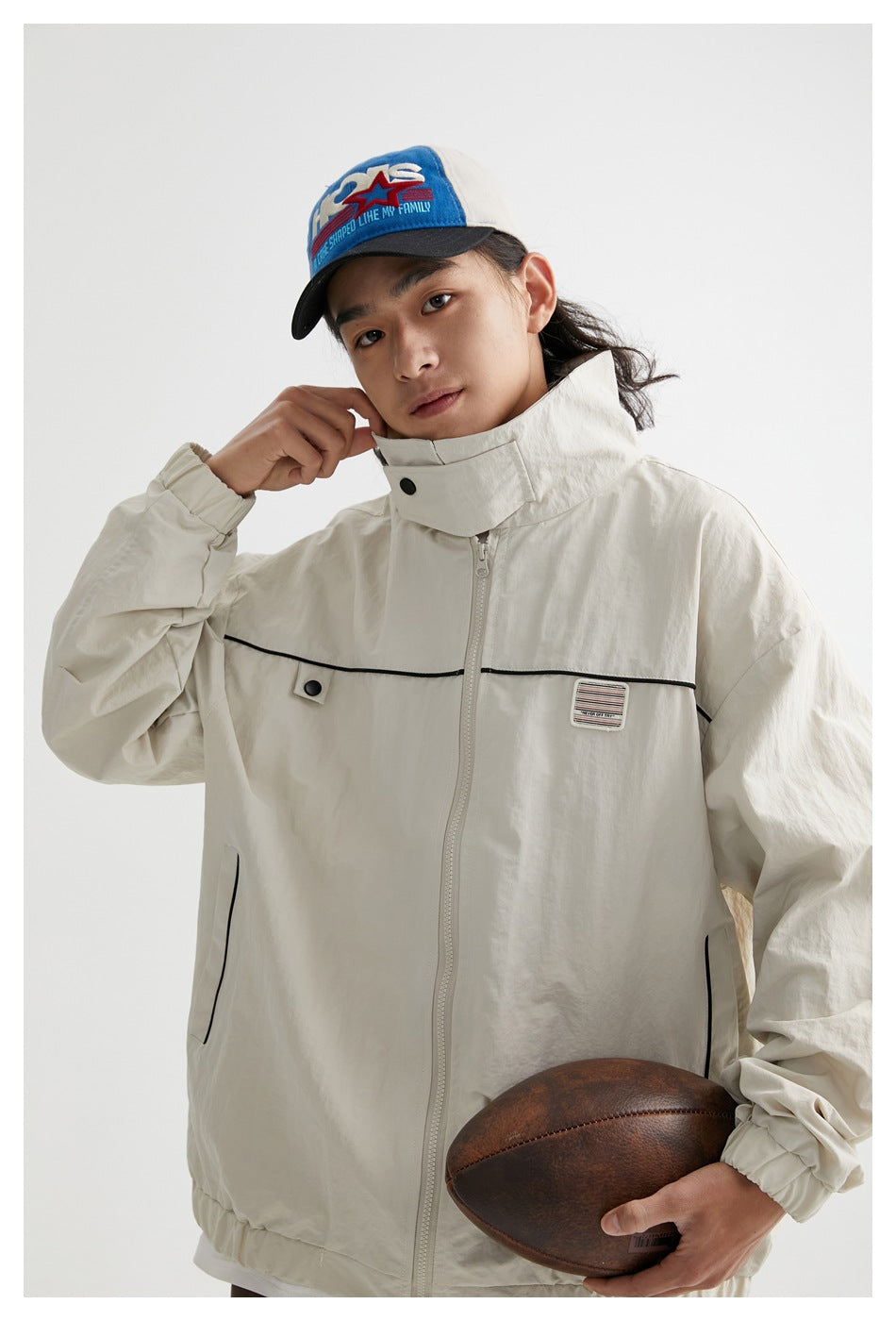 Close view of a model in an ivory high-collar jacket with black piping and zip pockets, holding a football.