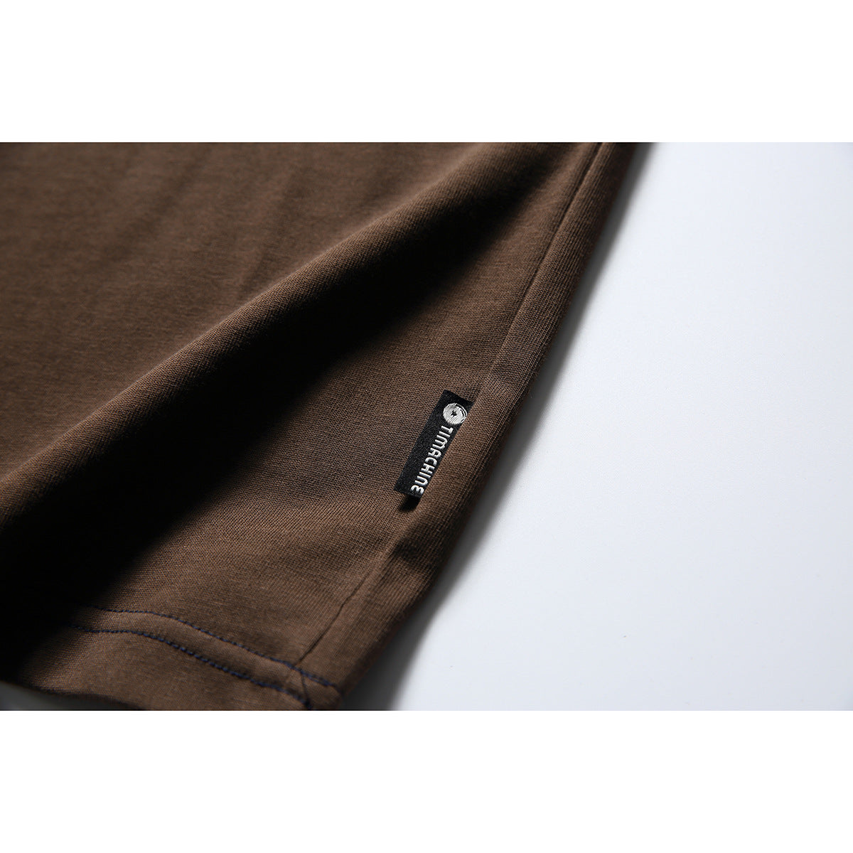 Close-up of the brown T-shirt side hem showing a small black woven label with a white logo.