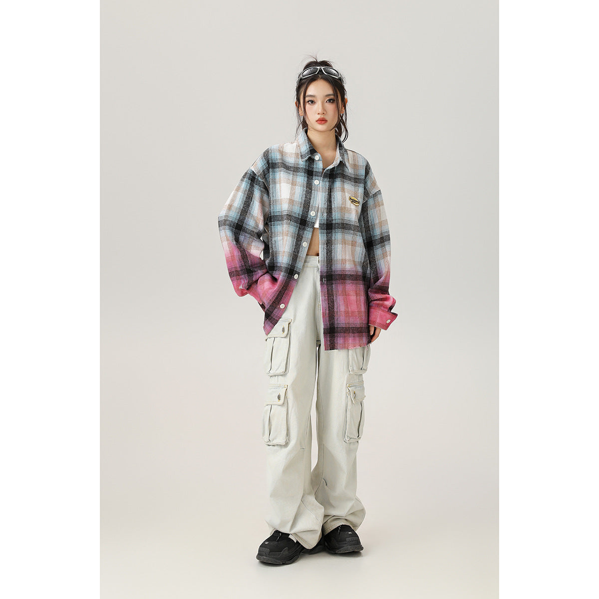 Full-length model wearing an oversized blue-gray plaid shirt fading to pink, light cargo pants, and black sneakers.