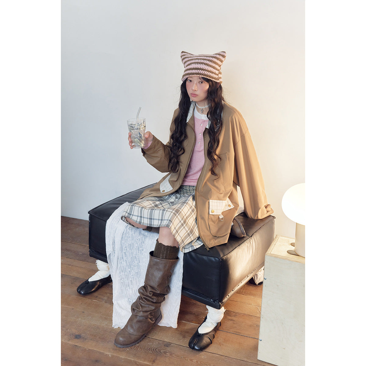 Seated model wears a brown jacket with plaid pocket details, a pink top, and a blue-gray plaid skirt.
