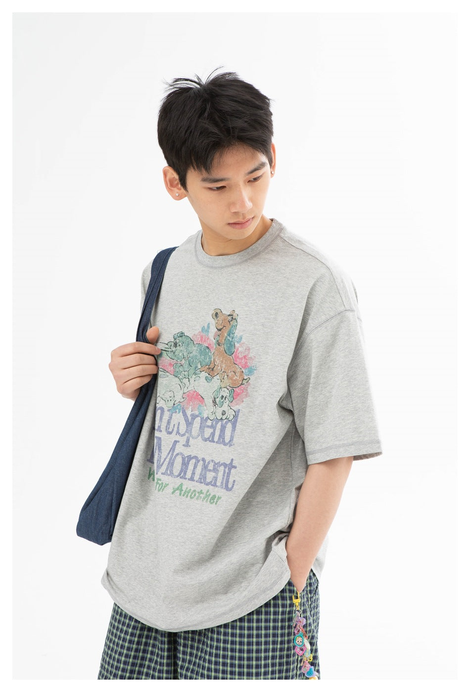 Model wearing a gray graphic T-shirt with illustrated animals and green plaid shorts, holding a navy shoulder bag.