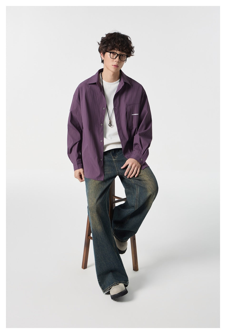 Model seated on a stool wearing a muted purple button-front shirt, white tee, and wide-leg faded jeans.