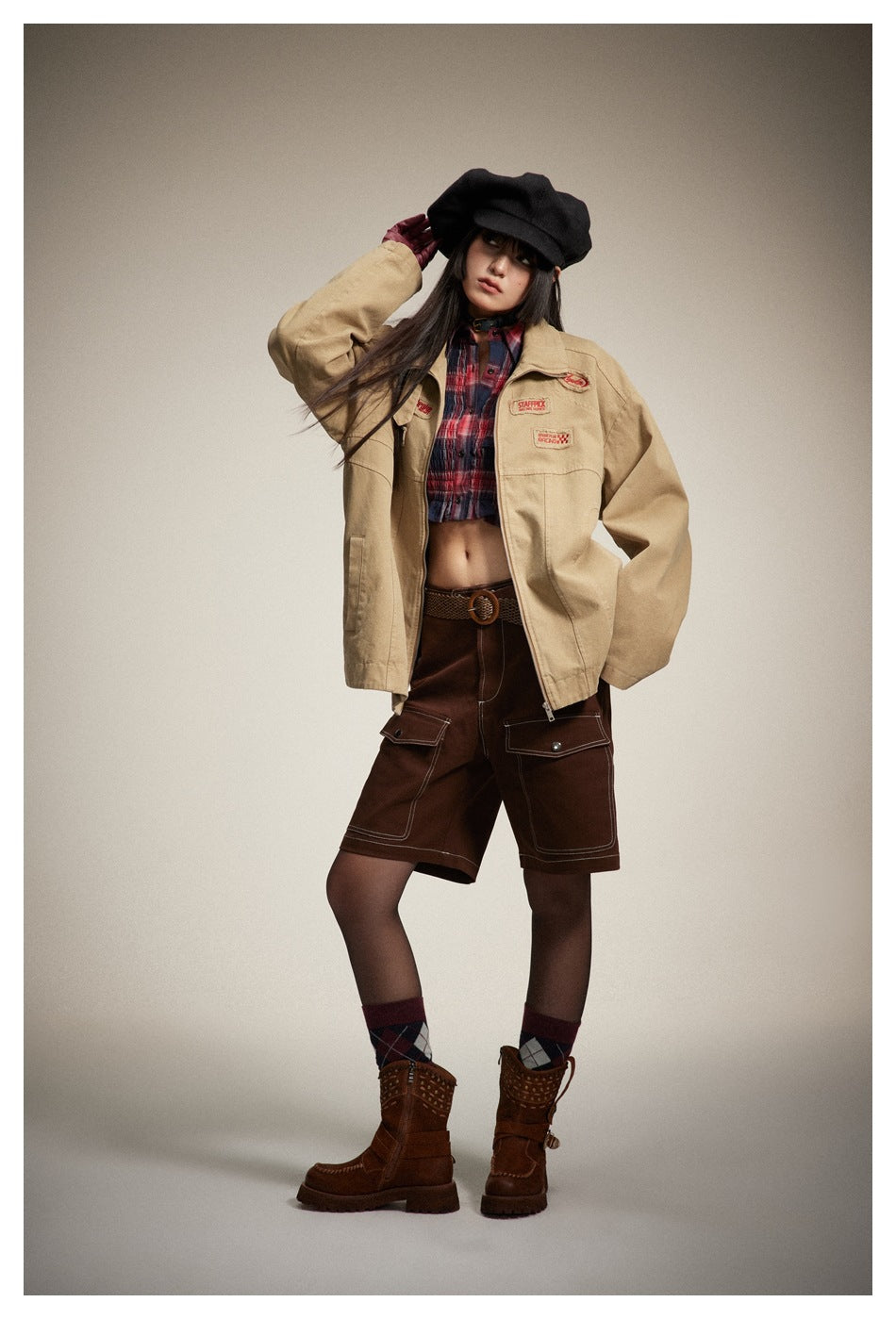 Model wearing an open khaki jacket with chest patches over a plaid top, brown shorts, black cap and brown boots