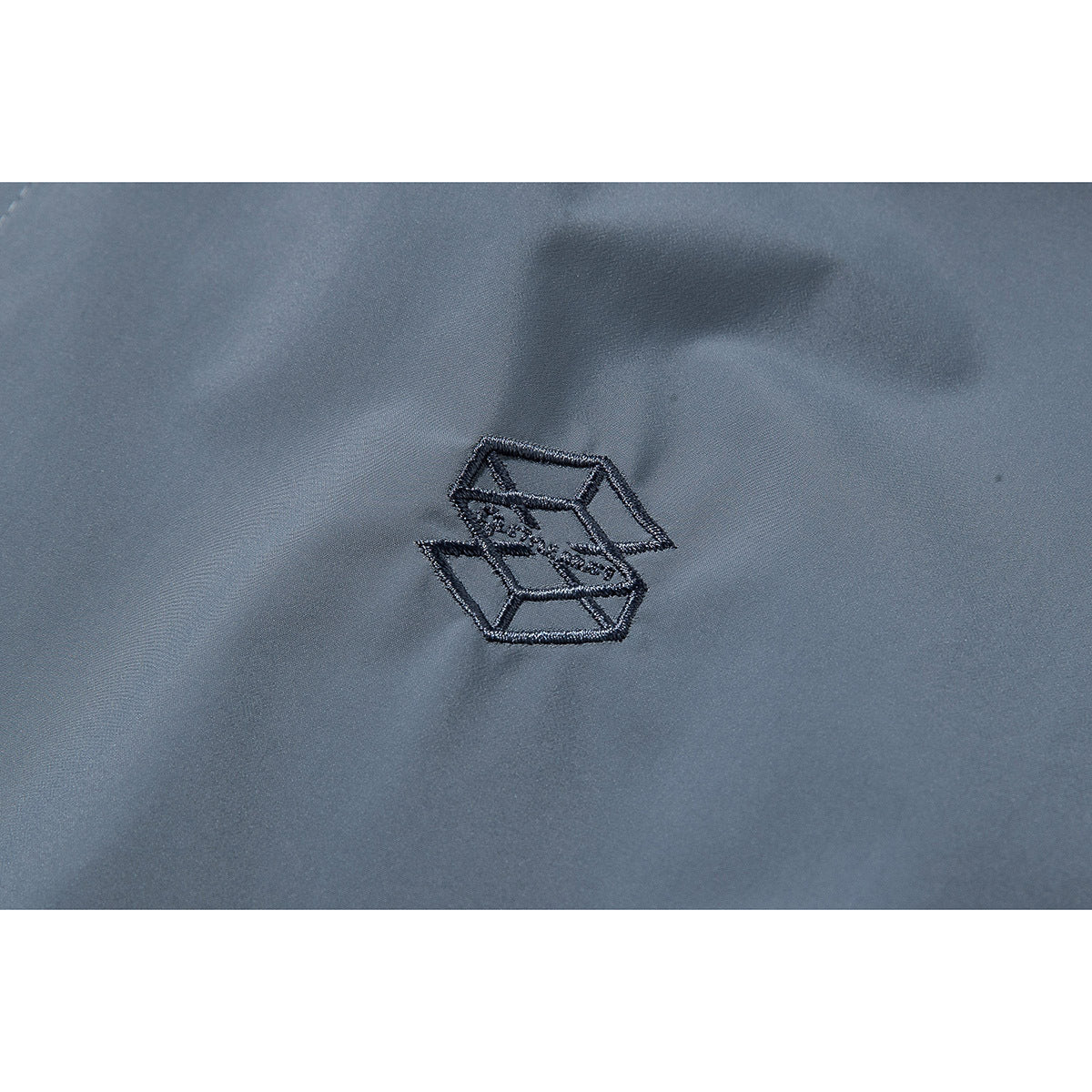 Close-up of a small dark embroidered geometric emblem on slate-blue jacket fabric.