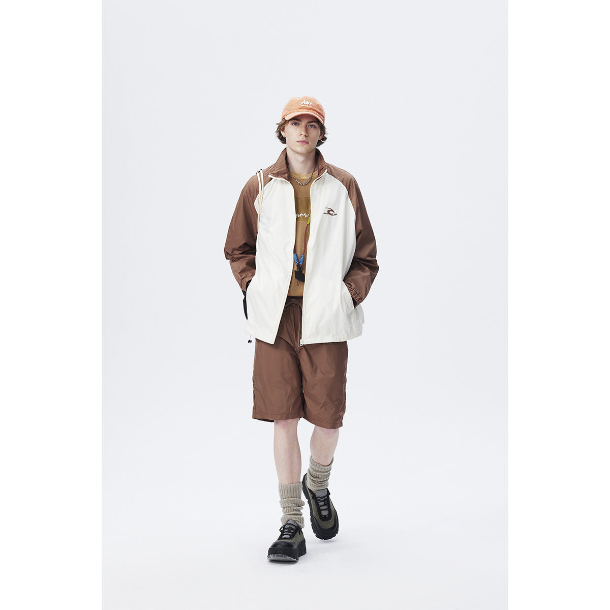 Model wearing a white jacket with brown sleeves, brown shorts, and an orange cap.