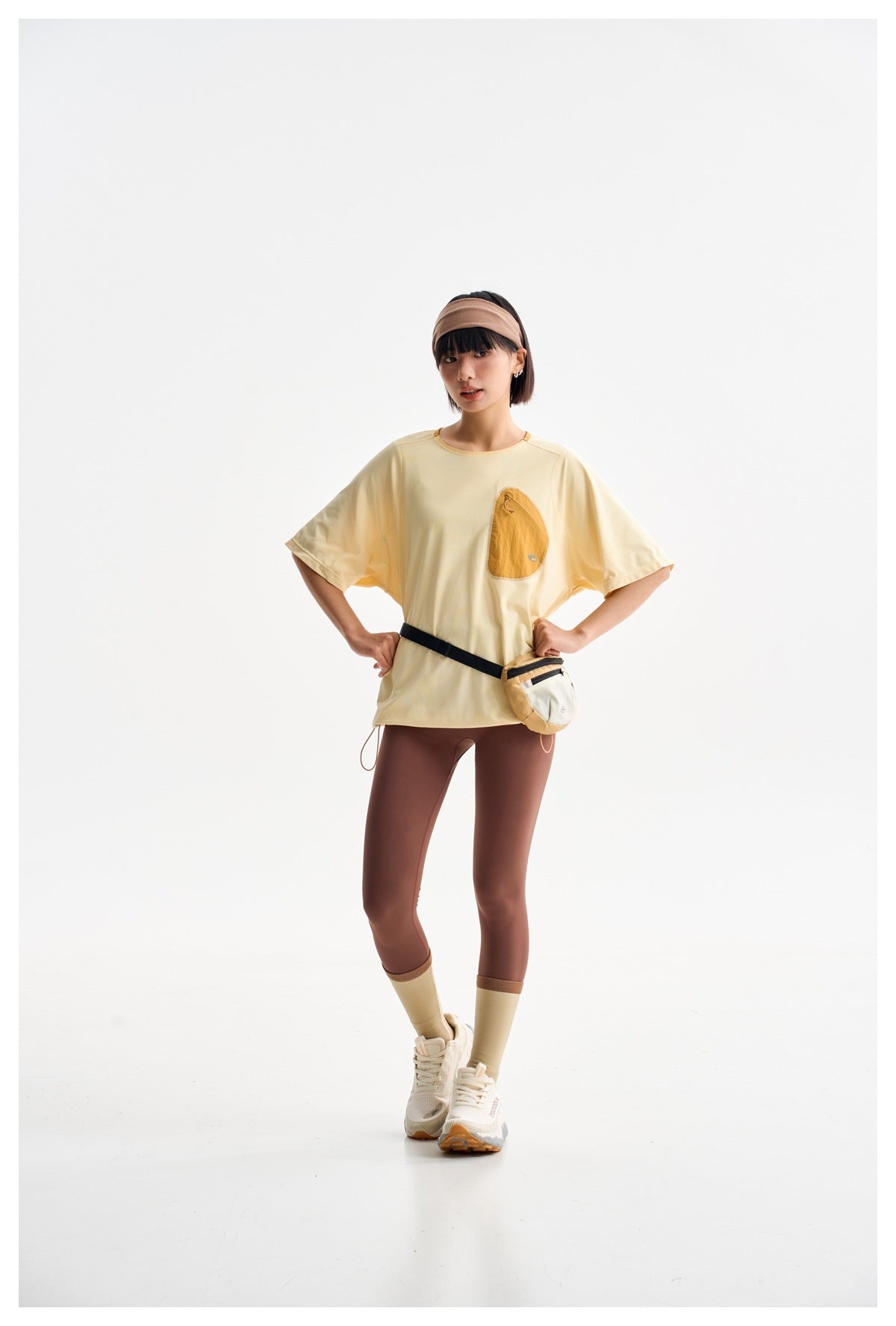 Model wearing a pale yellow short-sleeve top with a mustard chest pocket, brown leggings, and a waist pouch.