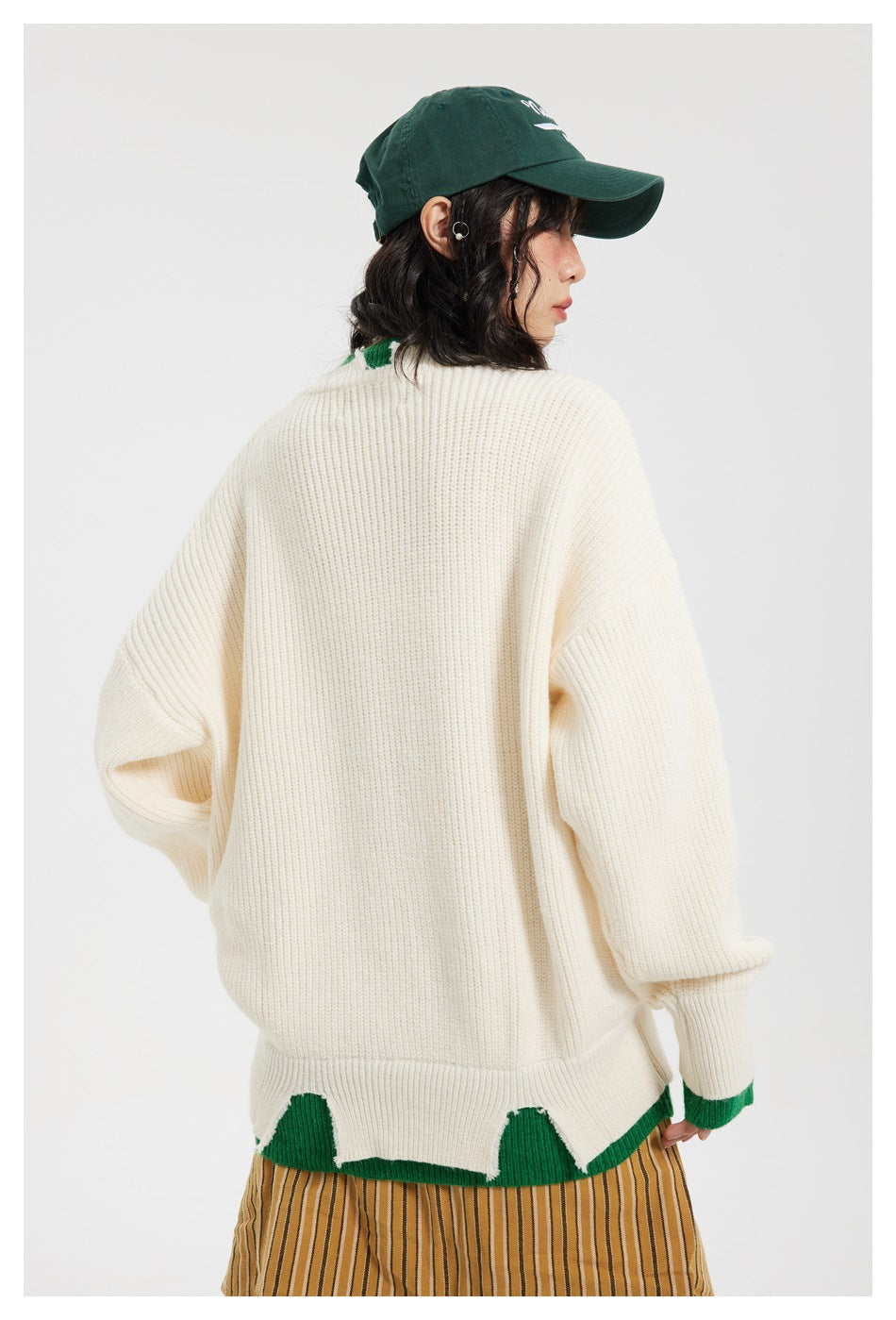 Model shown from behind in an ivory ribbed sweater with green trim and an uneven hem, wearing a dark green cap.