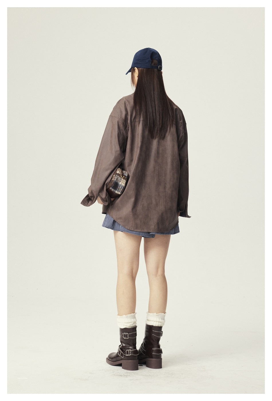 Rear view of a model wearing an oversized brown button-up jacket, checked shorts, navy cap, white socks and dark boots.