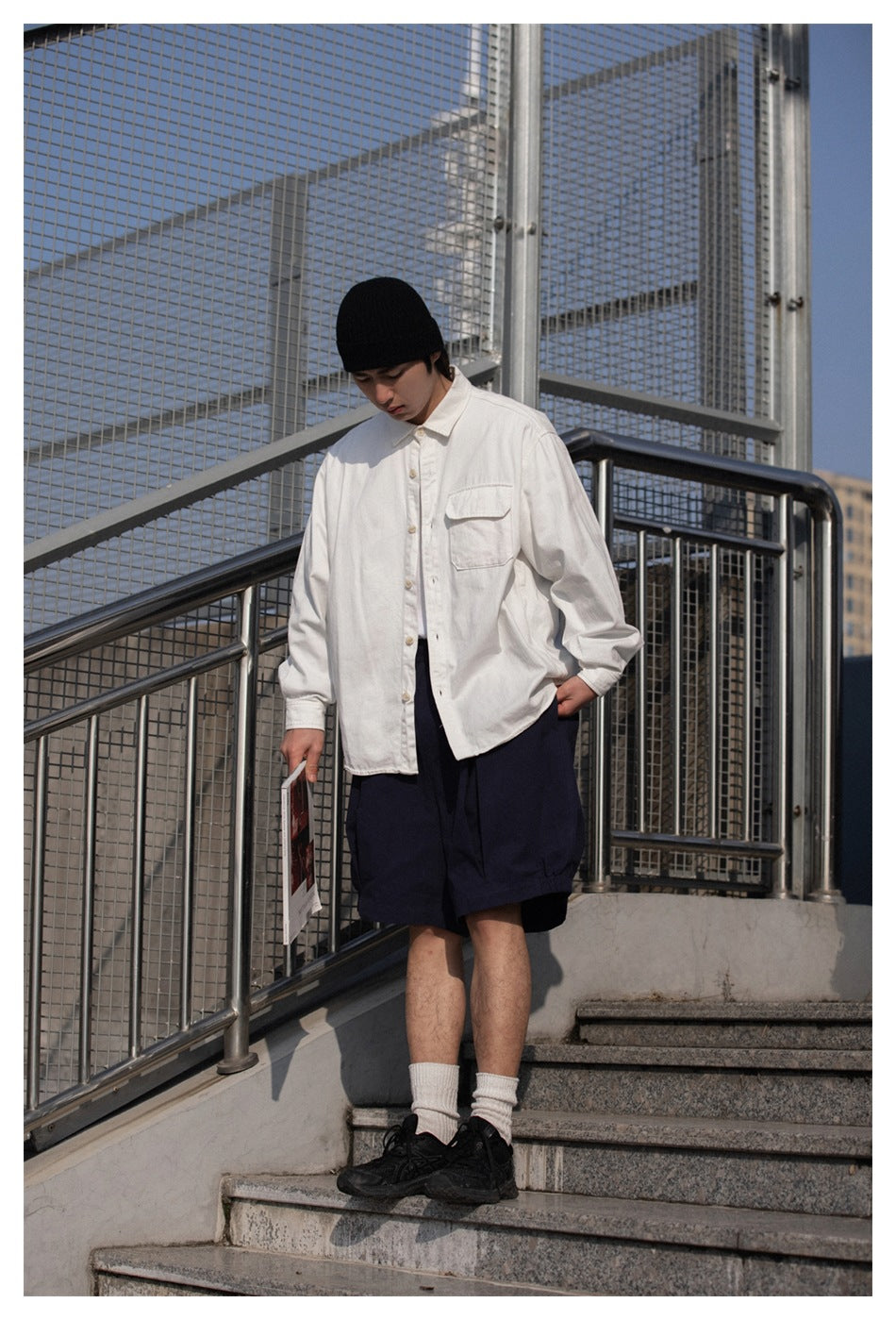 Model wearing loose navy shorts with a white button-front shirt and chest pocket, standing on outdoor steps.