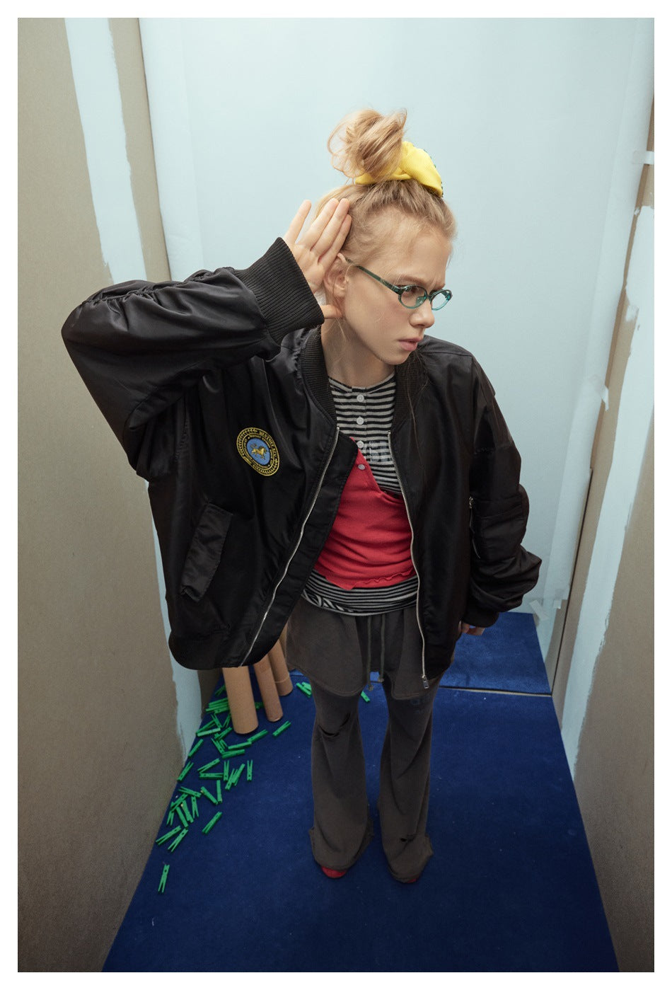 Model wearing an open black bomber jacket with a gold-and-blue chest patch, ribbed trim, and zip pockets.