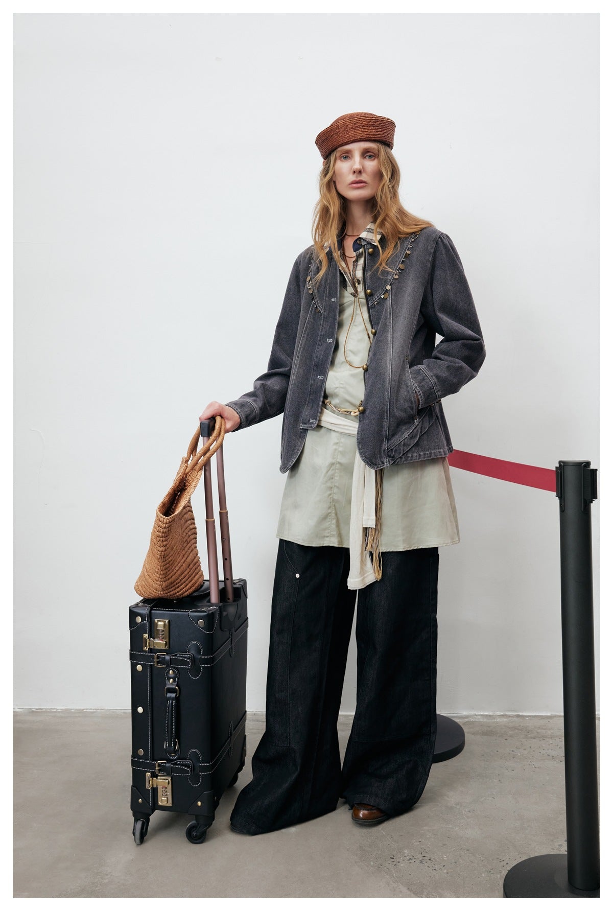 Model in the gray denim jacket with a hand in pocket, standing beside a black suitcase and holding a straw bag