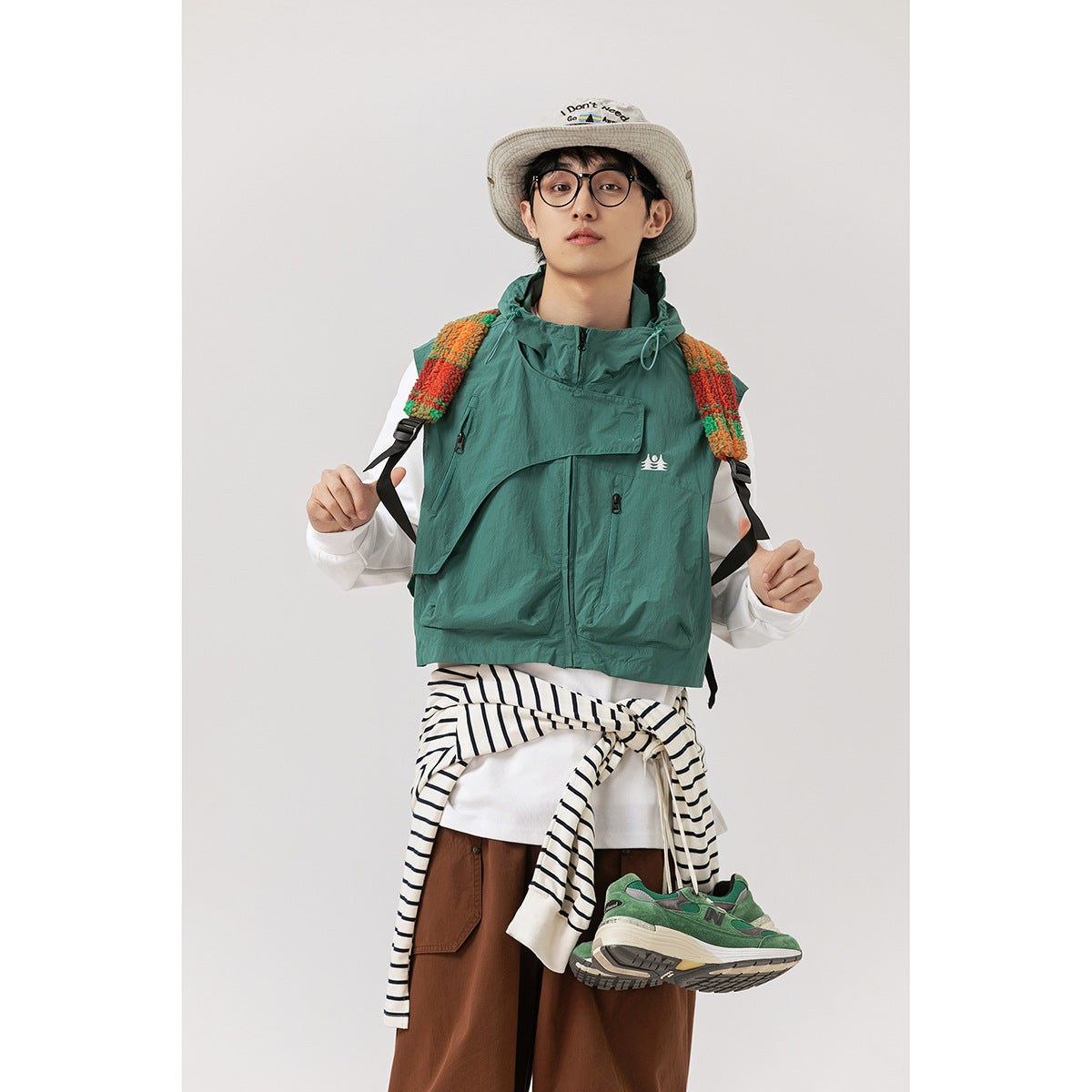 Model wearing a teal hooded utility vest with front pockets over a white shirt, styled with a bucket hat and brown pants.