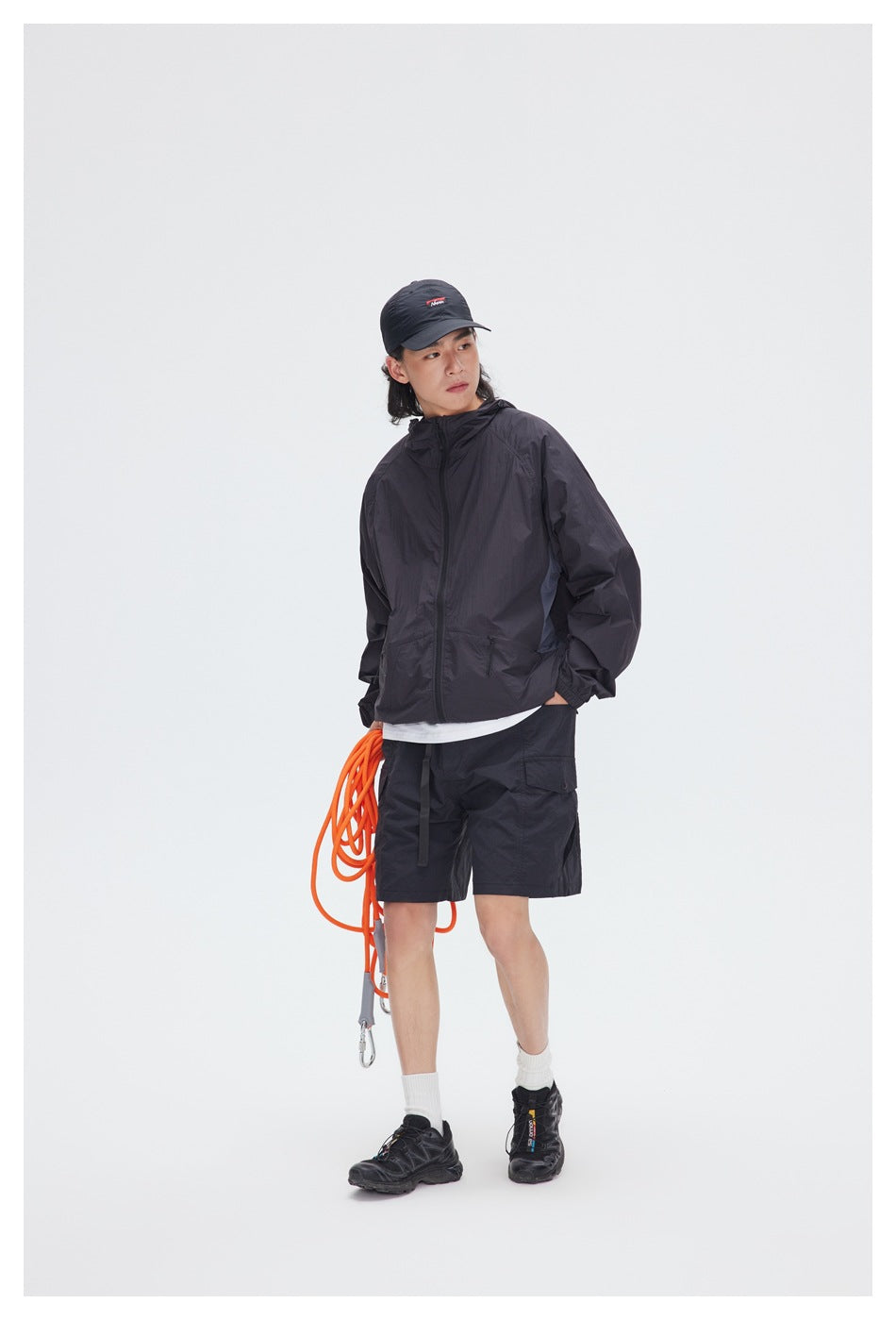 Model wearing black hooded zip jacket over a white tee with black cargo shorts, cap and sneakers
