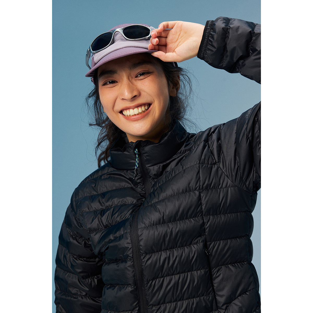 Smiling model in a black quilted zip jacket and lavender cap with sunglasses perched on the brim.