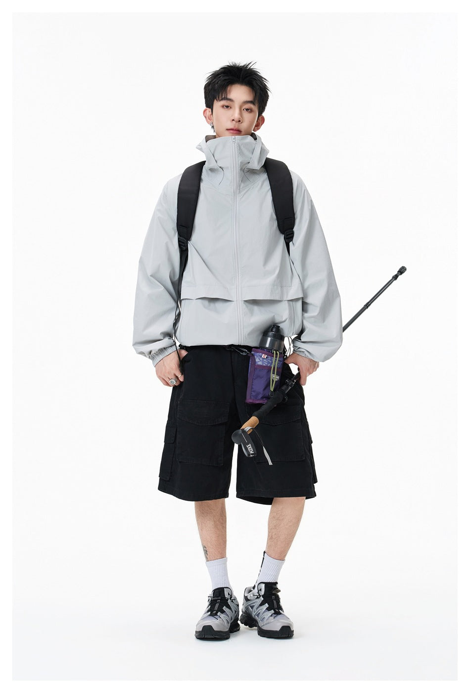 Full-length model wearing a light gray high-collar zip jacket and black cargo shorts with a backpack.