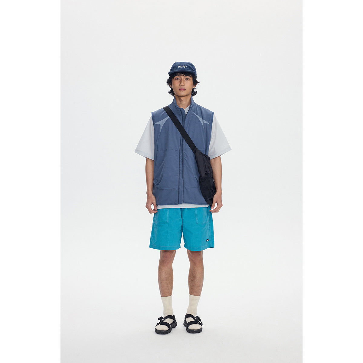 Front view of a model wearing a slate-blue zip vest over a white T-shirt with turquoise shorts and a black shoulder bag.