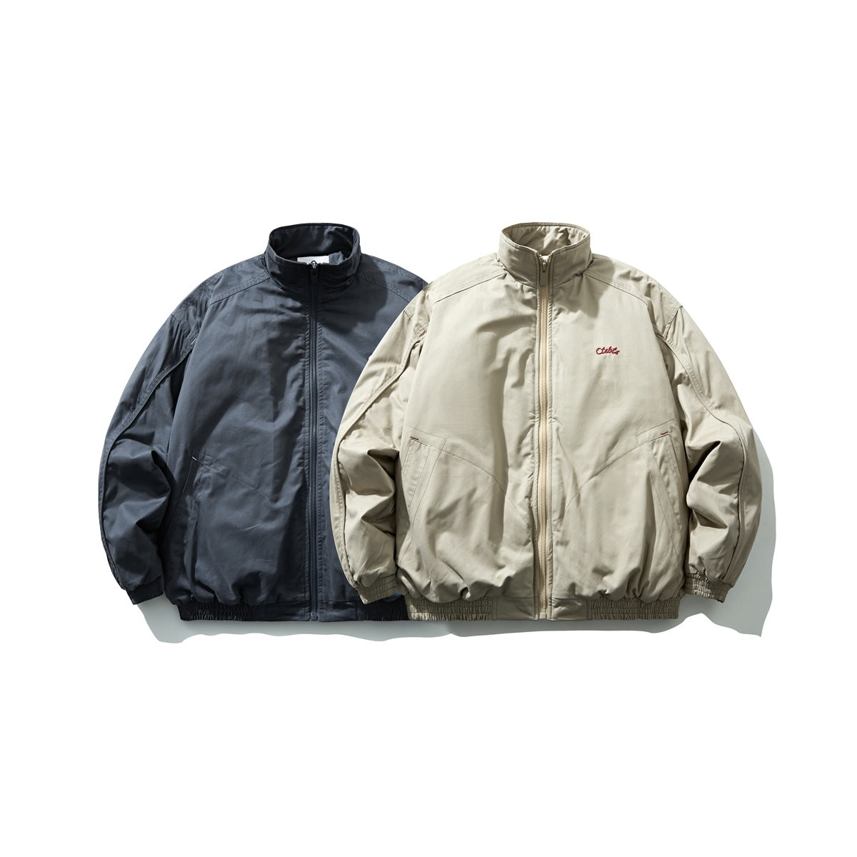 Two front-facing stand-collar zip jackets shown together in charcoal blue and light beige.