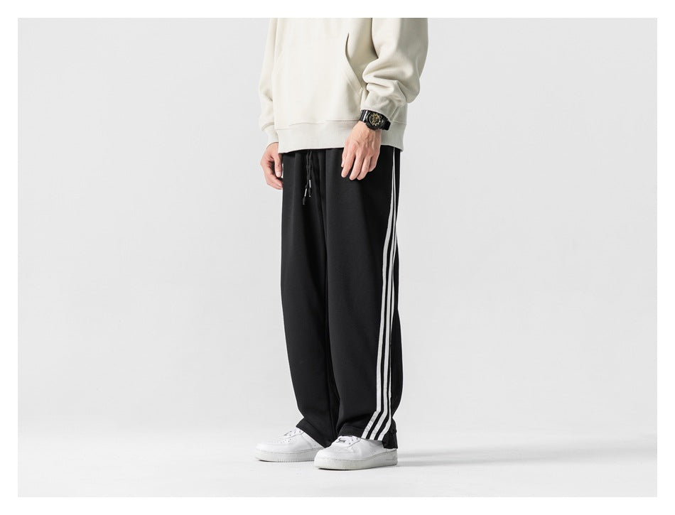 Cropped model in a front view wearing loose black track pants with three white side stripes.